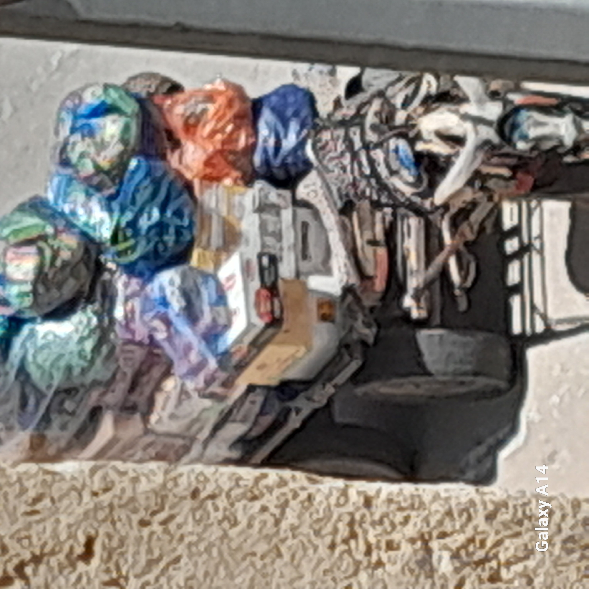 In Khan Yunis, Palestinian Territories, a motorbike is laden with a large, precarious stack of goods, predominantly in colorful plastic bags and boxes. The motorbike itself is mostly obscured by the load. The items are piled high, reaching significantly above the rider's seat, and appear to be various consumer products. The scene suggests a mobile vendor or someone transporting a substantial amount of merchandise. The background shows a textured wall and a light-colored surface, potentially a street or sidewalk. The overall impression is one of resourcefulness and necessity in a bustling urban or semi-urban environment.