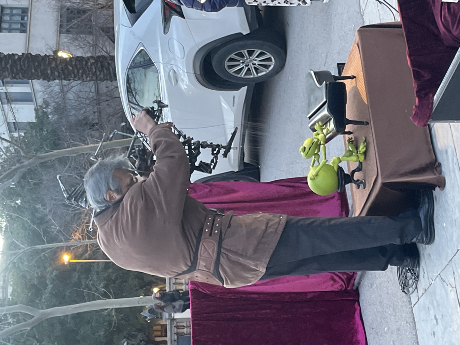 An older man with gray hair, wearing a brown jacket, dark pants, and a studded belt, stands in front of a table covered in a brown cloth. On the table are green frog figurines and a small black piano. Behind him is a white car. A deep purple velvet cloth hangs from the front of the table, reaching the ground. Trees and buildings are visible in the background.