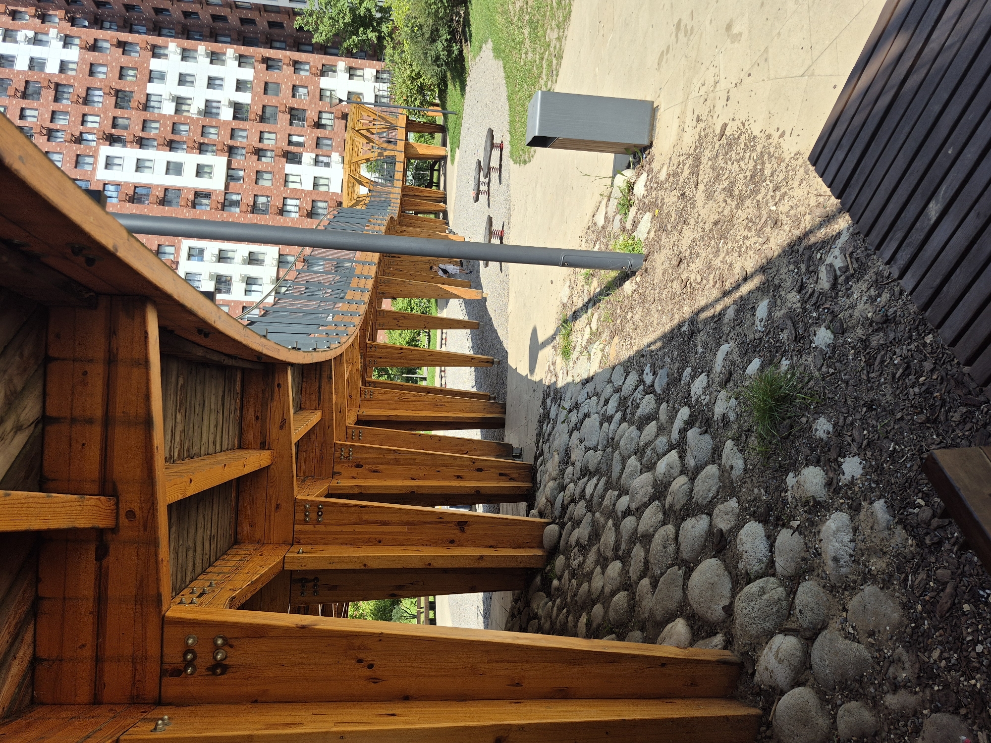 The image depicts a sunlit outdoor area featuring a distinctive wooden structure. This structure, with its warm wooden beams and metallic accents, forms a covered walkway or pavilion, supported by numerous angled wooden posts. The ground beneath the structure is a mix of large, smooth, grey stones and dark wood chips. Beyond the wooden construction, a modern residential building with a brick facade and numerous windows is visible. The area appears to be a park or public space, with green grass, trees, a path, a trash can, and what looks like playground equipment (spring riders). A person, dressed in light clothing and carrying a backpack, is walking on a path in the distance. The scene is bright and appears to be a warm day. This location is likely the "Khodynskoye Pole" park in Moskva, Russia, known for its contemporary architecture and recreational facilities.
