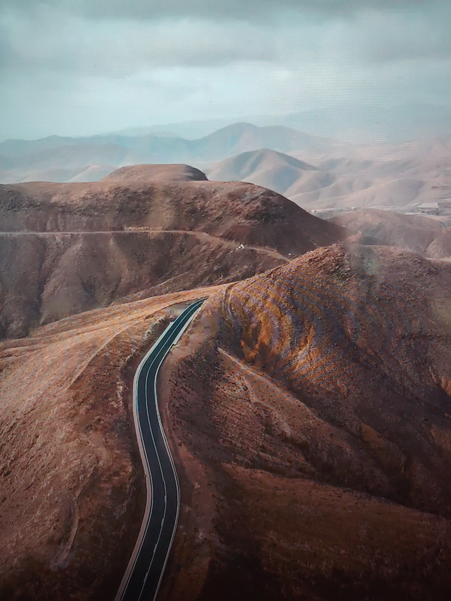 The image shows an aerial view of a winding road traversing a mountainous, arid landscape. The road, paved and marked with white lines, cuts through reddish-brown hills and mountains. In the background, a hazy, overcast sky is visible, with more distant mountains fading into the horizon. The environment is dry and barren, suggesting a desert or semi-arid region. There are no people, subjects, objects, actions, or interactions observed other than the road itself.