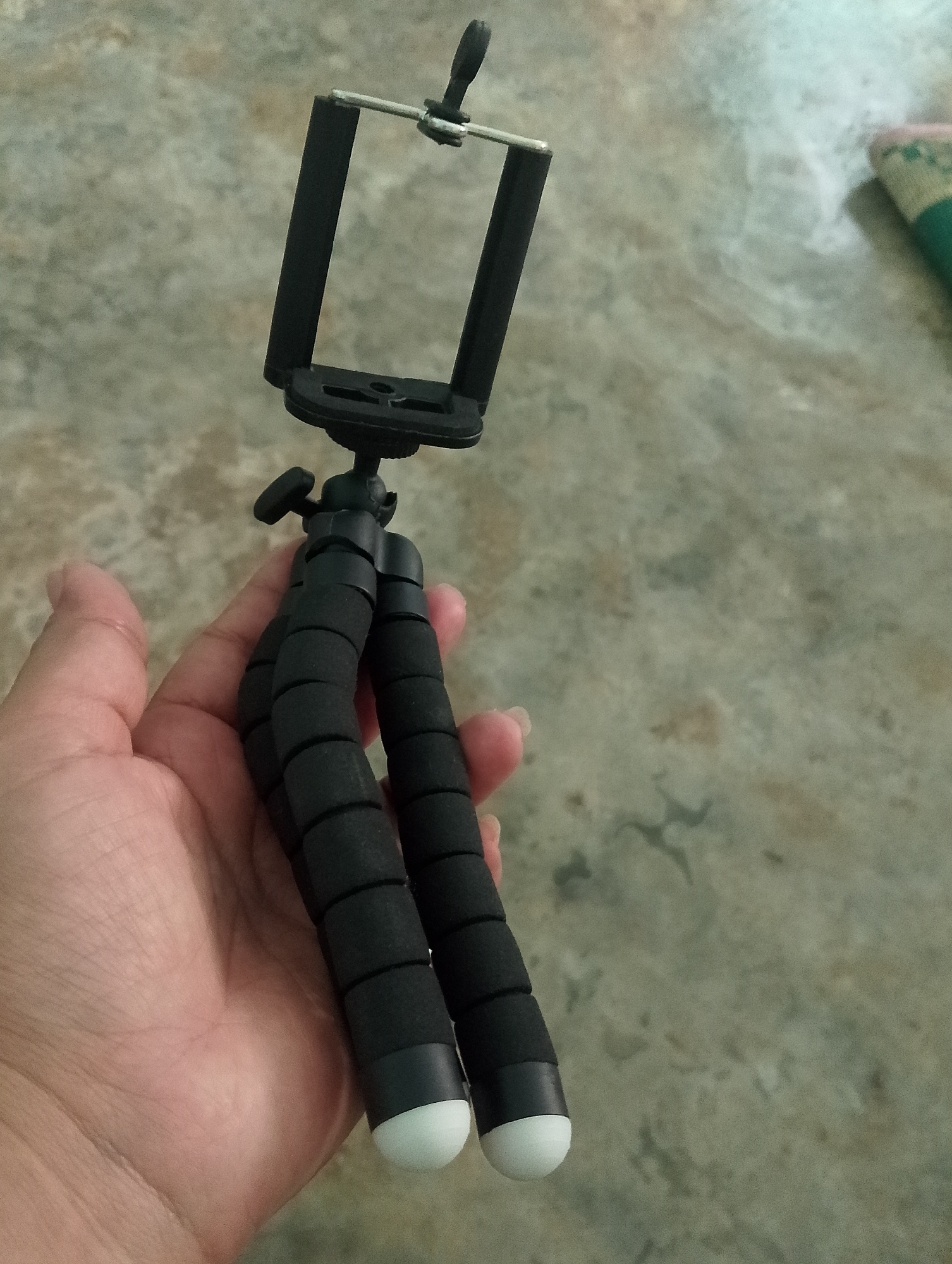 A hand is holding a black, flexible tripod with a phone holder attached. The tripod has two segmented, foam-covered legs with white caps at the bottom. The phone holder is black and designed to grip a mobile device. The background is a textured, gray floor with some lighter and darker patches. There is a hint of a green and pink object in the upper right corner, possibly a mat or towel. The image appears to have been taken indoors, given the floor texture. No specific details about the location in Serbelawan, Indonesia can be determined from the image itself.