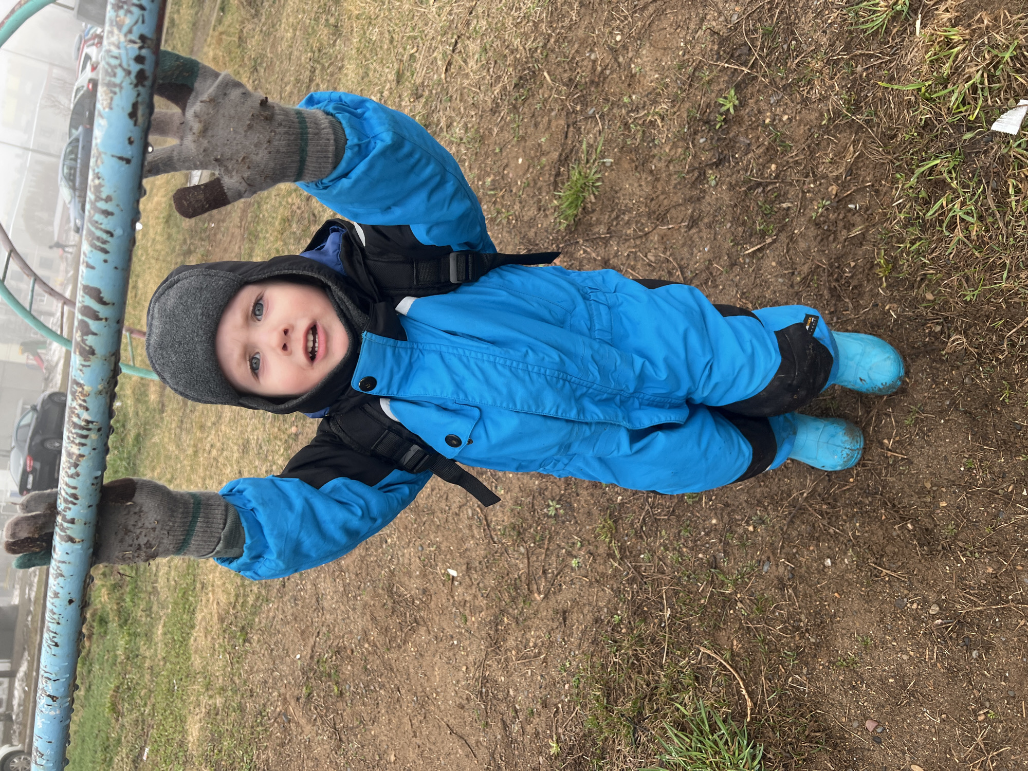 A young child, estimated to be between 2 and 4 years old, dressed in a bright blue snowsuit with black accents, a grey knit hat, and blue boots, is hanging from a blue metal playground structure. The child is wearing grey gloves that appear soiled with dirt or mud. The child is facing the camera with an open mouth and a visible gap in the upper front teeth.
The ground is a mix of dirt, patchy grass, and dead leaves. There is debris scattered on the ground. A car is visible in the background, partially obscured by fog or mist. The overall scene appears to be a playground area in a somewhat unkempt condition.
The location is assumed to be in the city of Марковский, Russia.