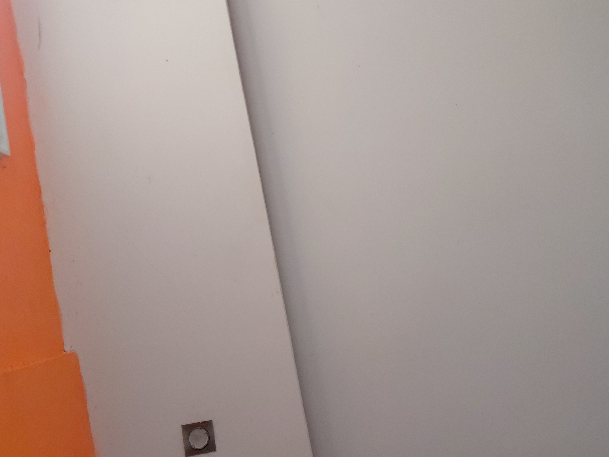 This image presents an abstract view of an interior space, focusing on wall surfaces and a small architectural detail. On the left side of the frame, a segment of a wall is painted in a striking, vibrant orange color. This orange wall meets a larger, expansive section of a wall painted in a light, neutral tone, resembling off-white or light grey. The intersection of these two walls forms a clear vertical line, indicating a corner or a change in wall plane.
Near the bottom-left corner of the frame, embedded within the light-colored wall and adjacent to the orange section, is a small, square-shaped metallic fixture. This fixture has a discernible circular element at its center, characteristic of a modern light switch, a button, or a low-profile electrical outlet.
The image is static, depicting architectural finishes with no human presence or activity. The surfaces appear smooth, indicating a finished interior. The lighting is diffused, making it impossible to determine the time of day or specific weather conditions. There is no visible text in the image. This scene is situated in Santa Ana, Venezuela.