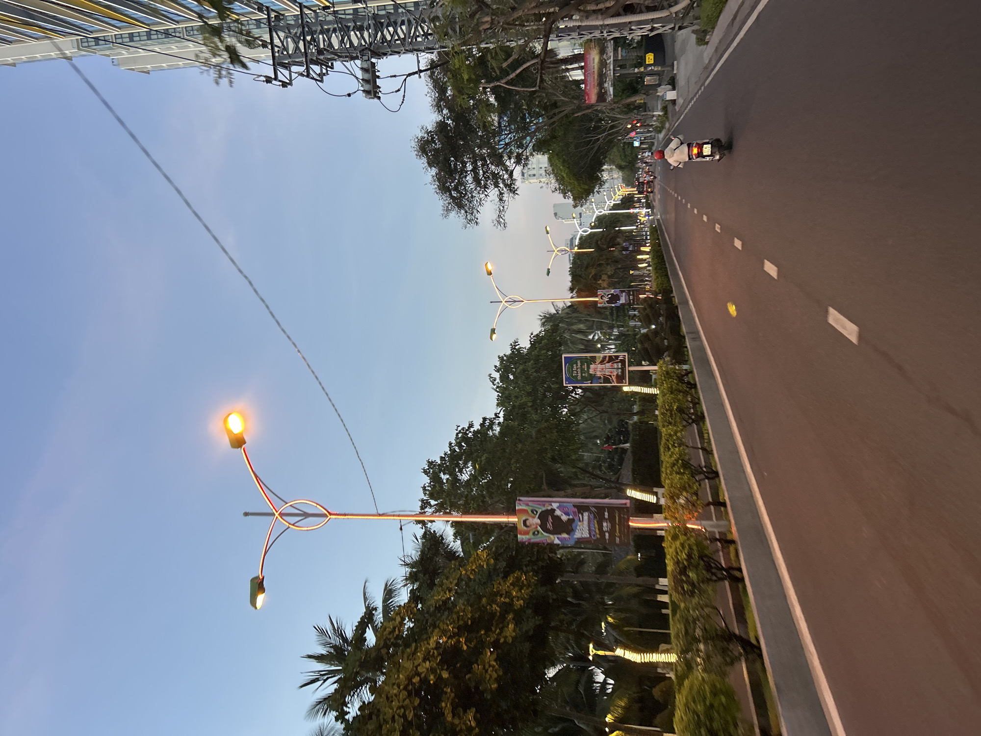 The image depicts a street scene in Nha Trang Ward, Vietnam, at dusk. A road with white dashed lane markings stretches into the distance, flanked by manicured bushes and trees on the left and a tall modern building with a metal utility pole on the right. Decorative streetlights with ornate poles and glowing lamps line the left side of the road. Several vertical banners are attached to these lampposts, featuring images of people and text. A lone individual on a scooter is traveling away from the camera down the road. The sky is clear with a gradient of blue and hints of orange from the setting sun.
