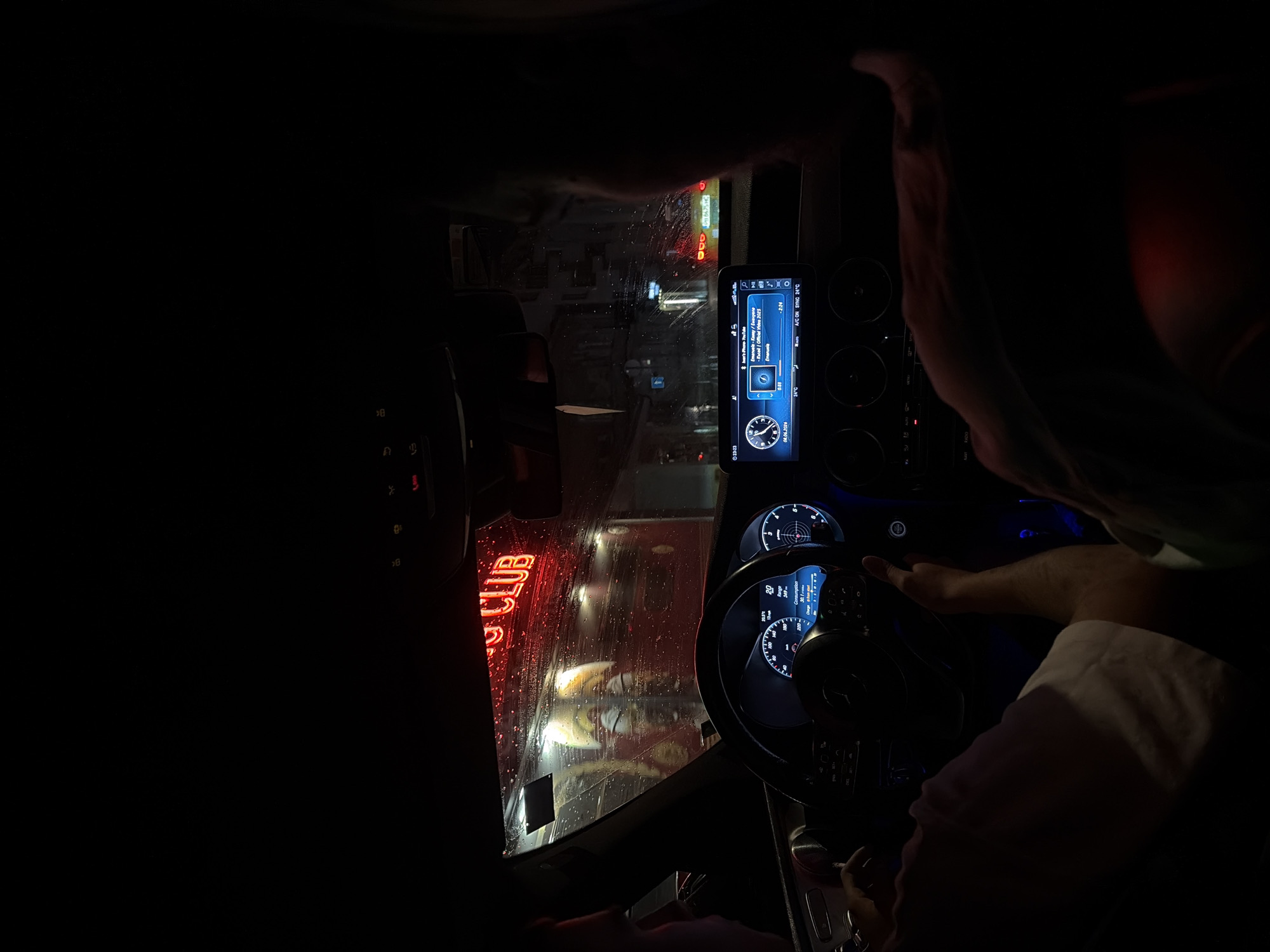 The image captures the interior of a Mercedes-Benz car at night, with a neon "CLUB" sign visible through the rain-streaked windshield. The car appears to be in motion, indicated by the blurred background. The dashboard displays illuminated instruments and a touchscreen infotainment system. Two individuals are visible within the car, one driving and another passenger. The driving individual's hands are on the steering wheel, and the passenger is partially visible in the foreground, looking away from the camera. The scene suggests an urban setting, possibly in Plovdiv, Bulgaria, given the neon sign and the overall atmosphere. The lighting is dim, with the neon sign and dashboard lights providing the primary illumination.