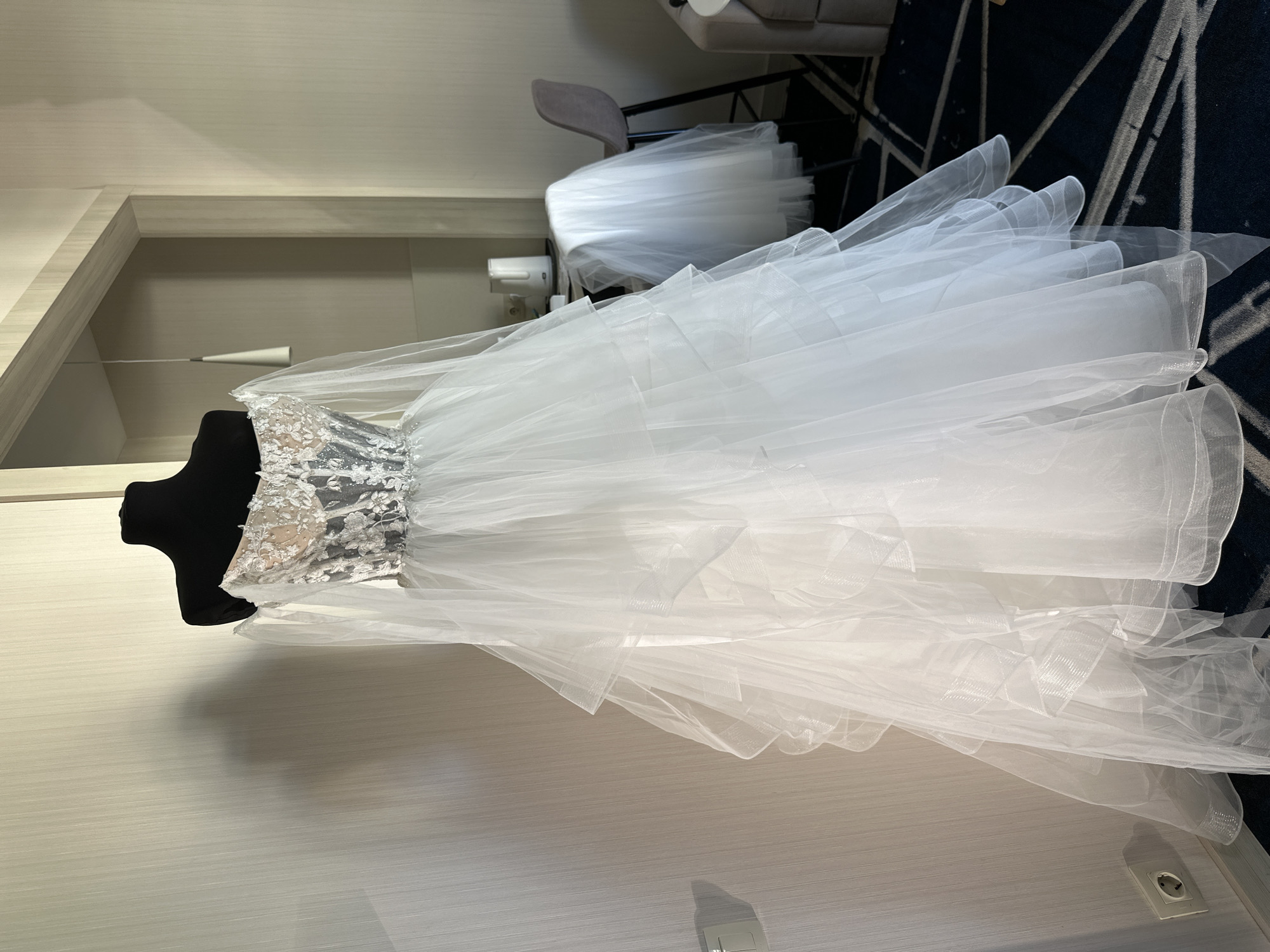 A white, tiered tulle wedding dress is displayed on a black mannequin in a well-lit room. The dress features an off-the-shoulder neckline with a sheer, embroidered bodice and long, flowing sleeves. The skirt is full and voluminous, with multiple layers of ruffled tulle. The background shows a wall with subtle vertical texturing, a doorway leading to another room, and a table with a kettle. A chair draped with a veil is visible in the background, along with a portion of a couch. The floor is covered in a dark carpet with a geometric pattern. The scene is devoid of any people.