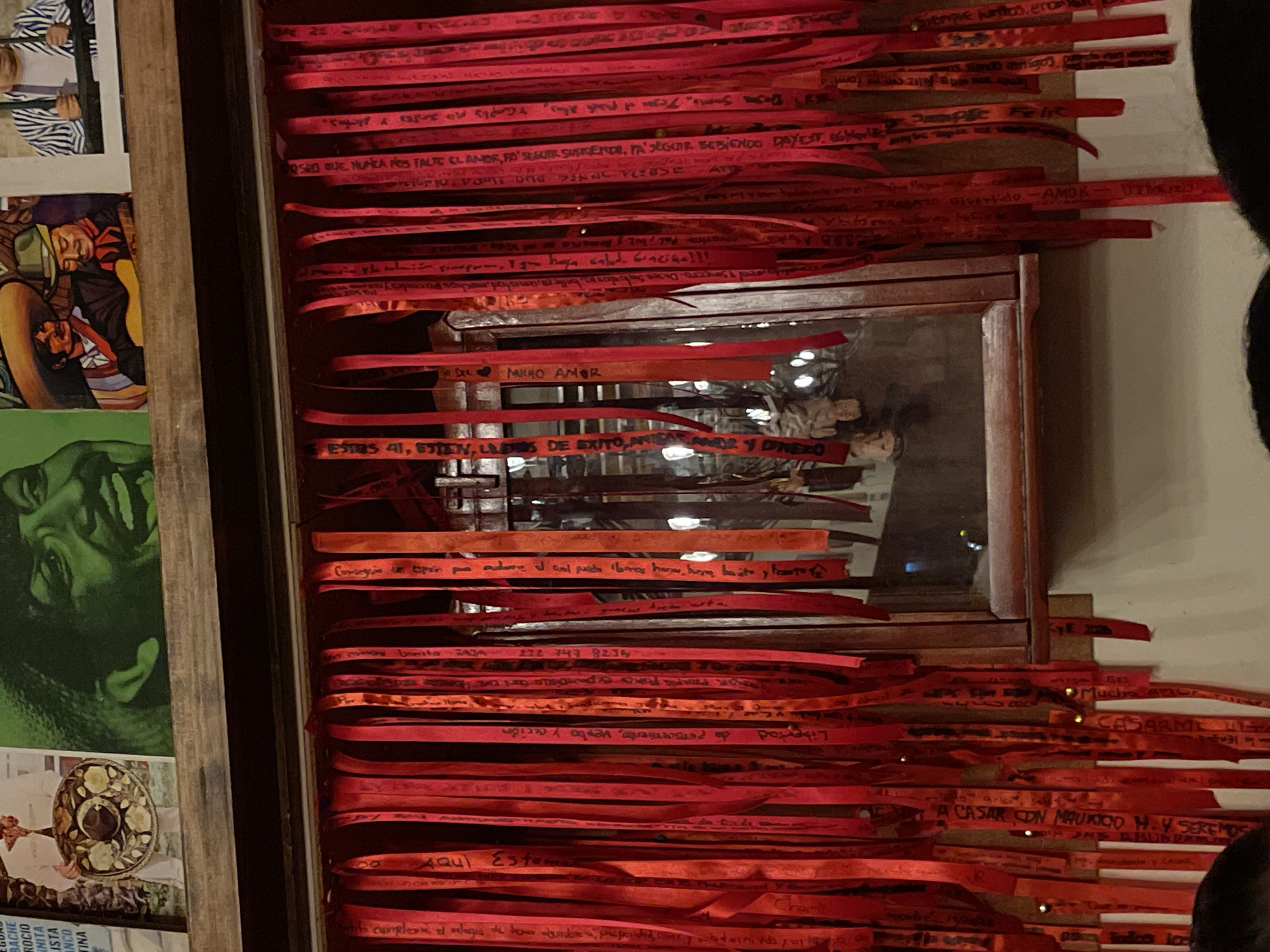 The image depicts a wooden framed altar covered in red ribbons with handwritten wishes, prayers, and dedications. The altar has a mirror in its center. To the left of the altar are other framed images and posters. The ribbons have messages written in Spanish, with phrases like "Mucho Amor" (Much Love), "Estar bien" (be well), and names and dates. The image seems to be shot indoors with warm, natural light. Based on the OCR text that includes references to "BACHE", "ROCIO", "INITA", "ISTA", "INCO" "TINA", the picture was taken at the "Santuario del Señor de la Preciosa Sangre", located in the historic center of Puebla, Mexico.