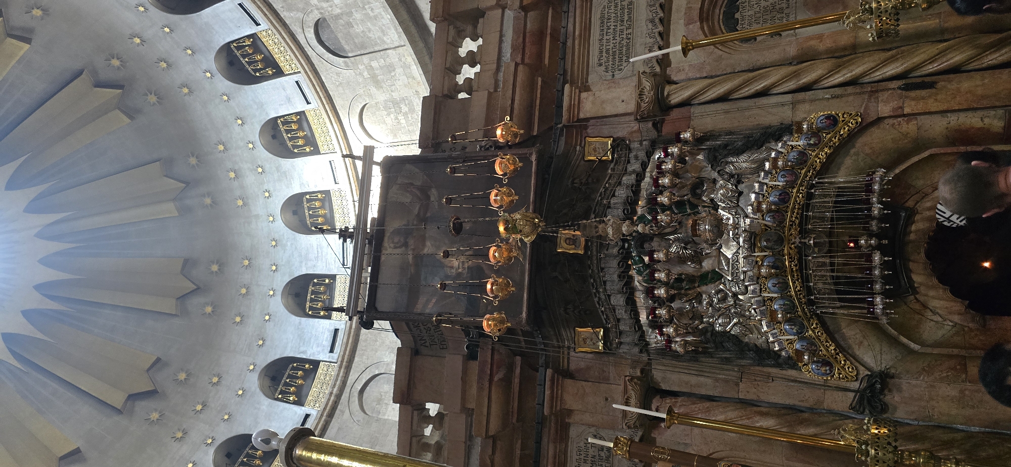 The image depicts the interior of the Church of the Holy Sepulchre in Jerusalem, Israel, a significant Christian pilgrimage site. The scene focuses on a section of the church's ornate interior, characterized by its religious architecture and decorative elements.
Dominating the upper part of the image is a large, domed ceiling, illuminated by light shafts from above. The dome is decorated with rows of golden star-like ornaments and features segmented arches. Below the dome, several arched niches are visible, each containing hanging lanterns.
In the mid-ground, a large, dark tapestry or painting hangs above an elaborate, multi-tiered structure. This structure appears to be a reliquary or altar, adorned with intricate silver sculptural figures, possibly depicting biblical scenes or religious motifs. Numerous smaller lamps, many with red-tinted glass, hang from chains above and around this central piece. Golden frames containing icons or small portraits are also affixed to the walls.
The lower section of the image shows architectural details such as stone columns, some with a twisted or rope-like design, and portions of walls inscribed with what appears to be text, likely in Greek or another ancient language. Several tall, golden candlesticks with white candles are positioned on either side of the central altar structure.
The foreground shows the heads of several individuals, suggesting the presence of visitors or worshippers within the church. Their presence indicates the active use of the space as a religious and tourist destination. The overall ambiance is one of reverence and historical significance, with a focus on the intricate craftsmanship and religious symbolism prevalent in such sacred spaces. The lighting, with sunbeams filtering through, adds to the atmospheric quality of the scene.
