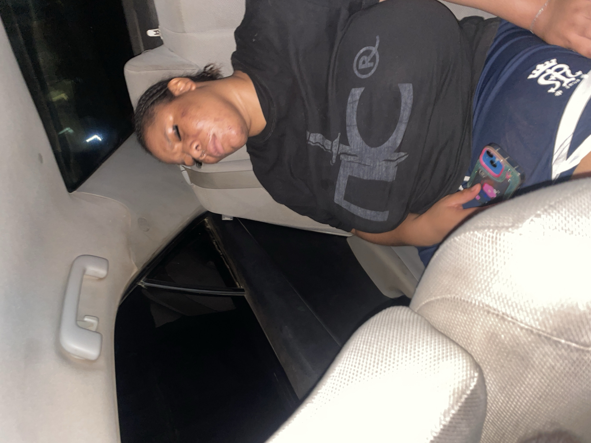 A medium shot captures a woman asleep in the back seat of a car, likely at night, given the darkness outside and the interior lighting. The car appears to be a sedan, with light-colored upholstery on the seats and a headliner with a grab handle. The woman is wearing a black t-shirt with a graphic print and navy blue shorts. Her eyes are closed, and her head is tilted to the side. A smartphone with a colorful case rests in her lap. The background is dark, suggesting the car is either parked or moving in a dimly lit area. The overall atmosphere is one of rest or exhaustion.