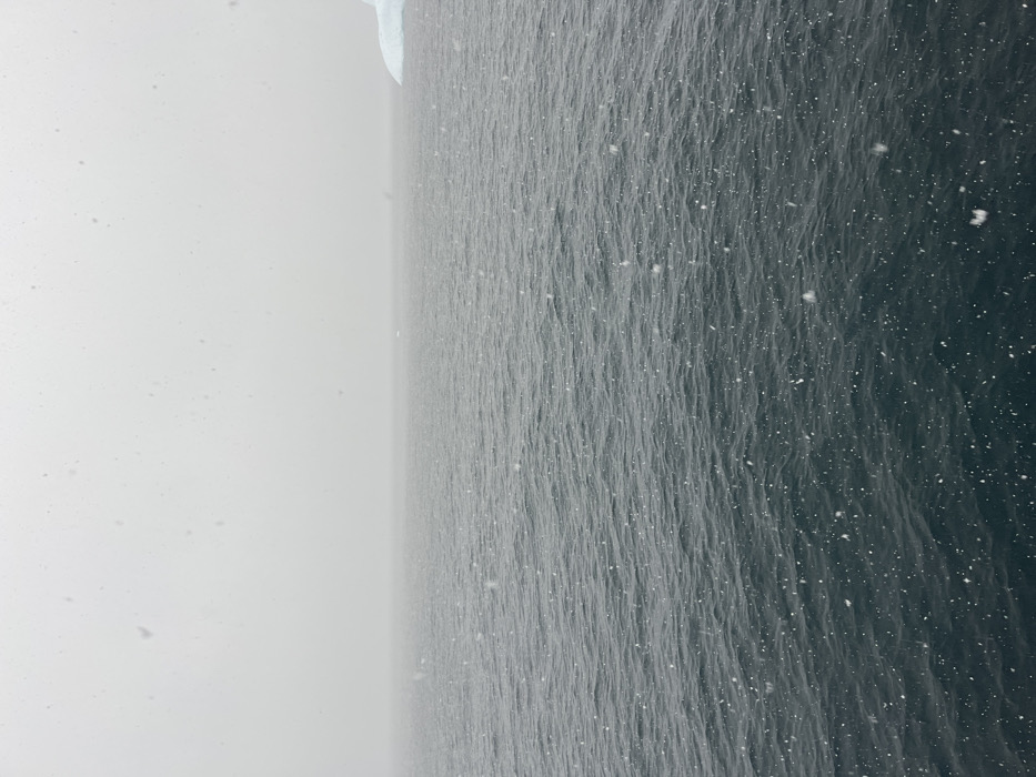 A sideways shot depicts a vast expanse of turbulent, dark water, possibly the ocean, under a snowfall. The left portion shows a hazy, almost white sky with visible snowflakes. The right side reveals the water's texture, small waves, and countless snowflakes dotting the surface. The upper left corner hints at a snowy, undefined edge, perhaps land or ice. The overall atmosphere is bleak and cold, evoking a remote, wintry scene.