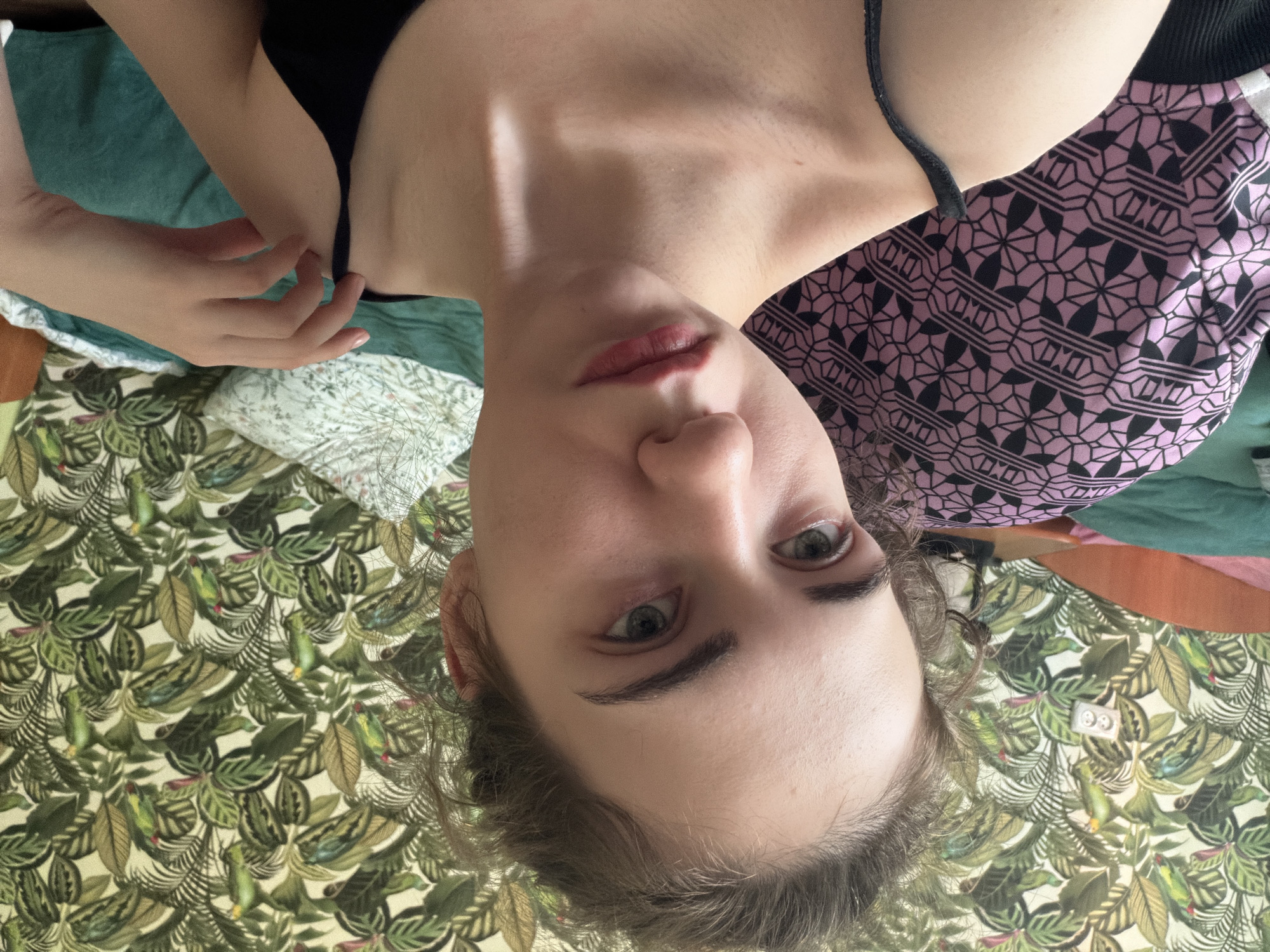 The image shows a close-up selfie of a young woman with light skin and brown hair. She is looking directly at the camera with a neutral expression. Her hair is pulled back loosely, and she is wearing a black spaghetti strap top. Her right hand is raised, and her fingers are gently touching her shoulder.
The background features a wall with tropical-themed wallpaper, featuring green leaves and colorful birds. A light-colored electrical outlet is visible on the wall. To the left of the woman, a patterned fabric, likely part of a blanket or pillow, with a pink and black geometric design is visible. There are also other pieces of fabric in shades of green and orange, suggesting the setting is indoors, possibly a bedroom or living room.
There is no indication of the location being in Zavodoukovsk, Russia.