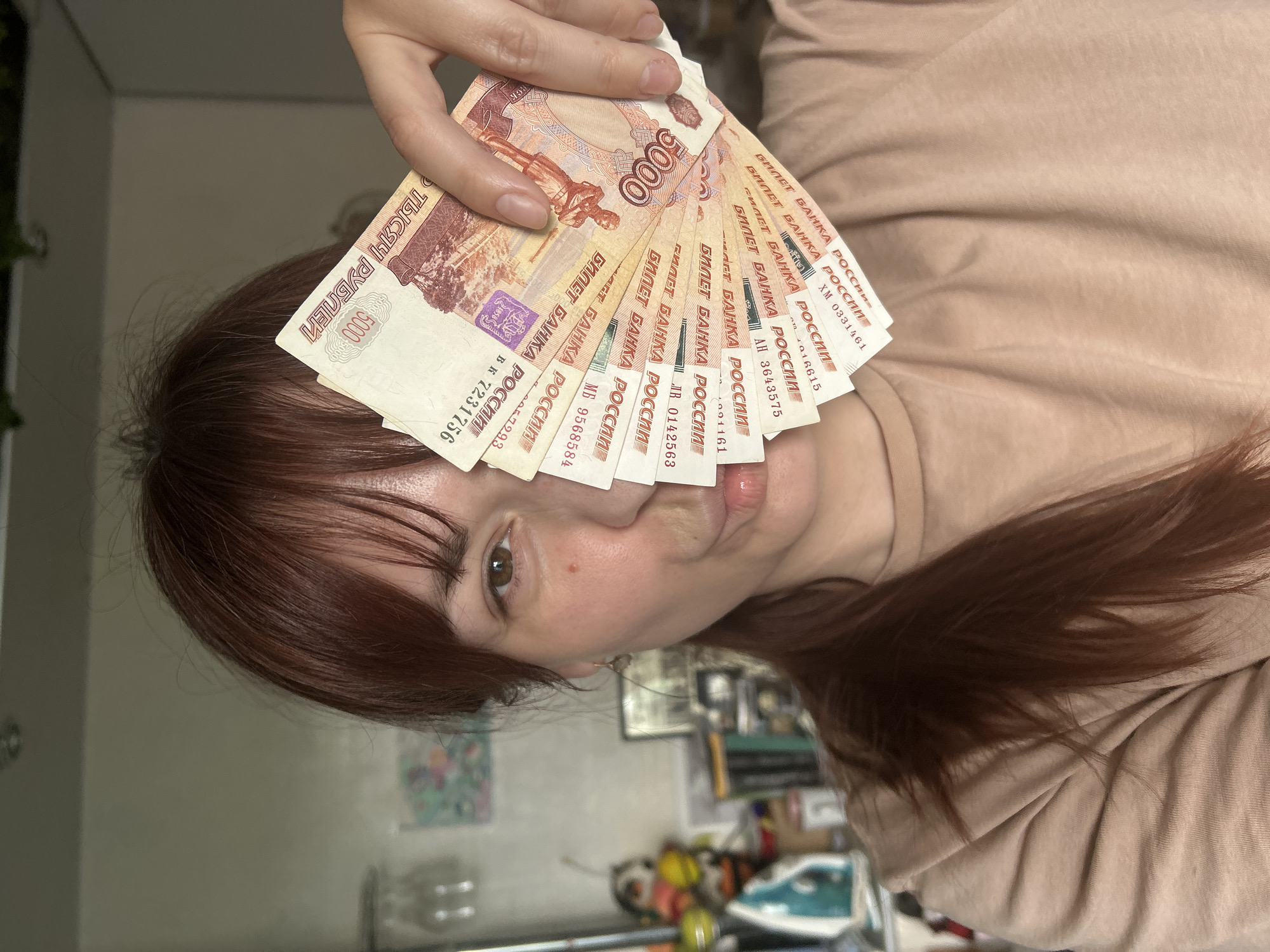 A woman is holding a fan of Russian 5000 Ruble banknotes in front of her face. The banknotes are predominantly reddish-orange in color. The location is not discernible from the image; there is no information to suggest it is in the city of Фоки, Russia.