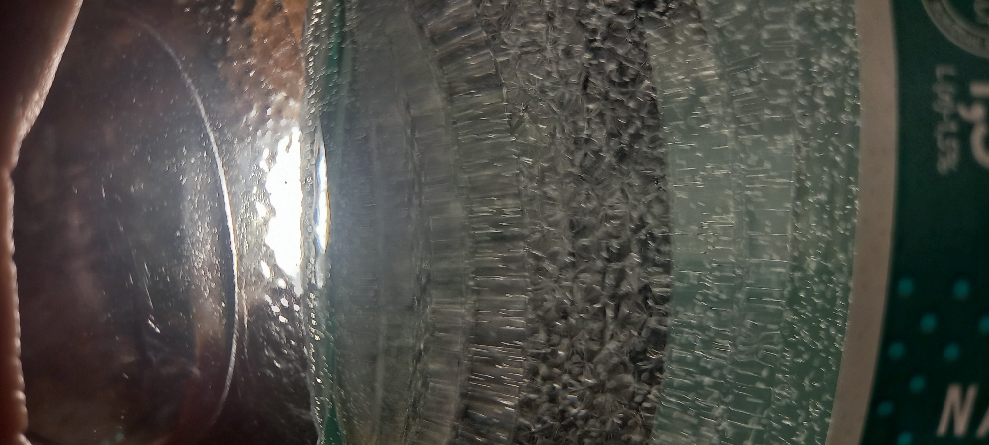 The image shows a close-up, distorted view of a clear glass bottle, possibly containing a beverage. The perspective is from inside the bottle, looking outwards. Light is refracting through the liquid and the glass, creating numerous bright highlights and patterns. The textured surface of the bottle's interior is visible, with what appear to be small bubbles or imperfections. The lower portion of the image shows a sliver of the bottle's label, with some green coloring and what looks like text or a logo. No specific location in Kryvyi Rih, Ukraine, can be determined from this image. There are no people or discernible actions taking place.