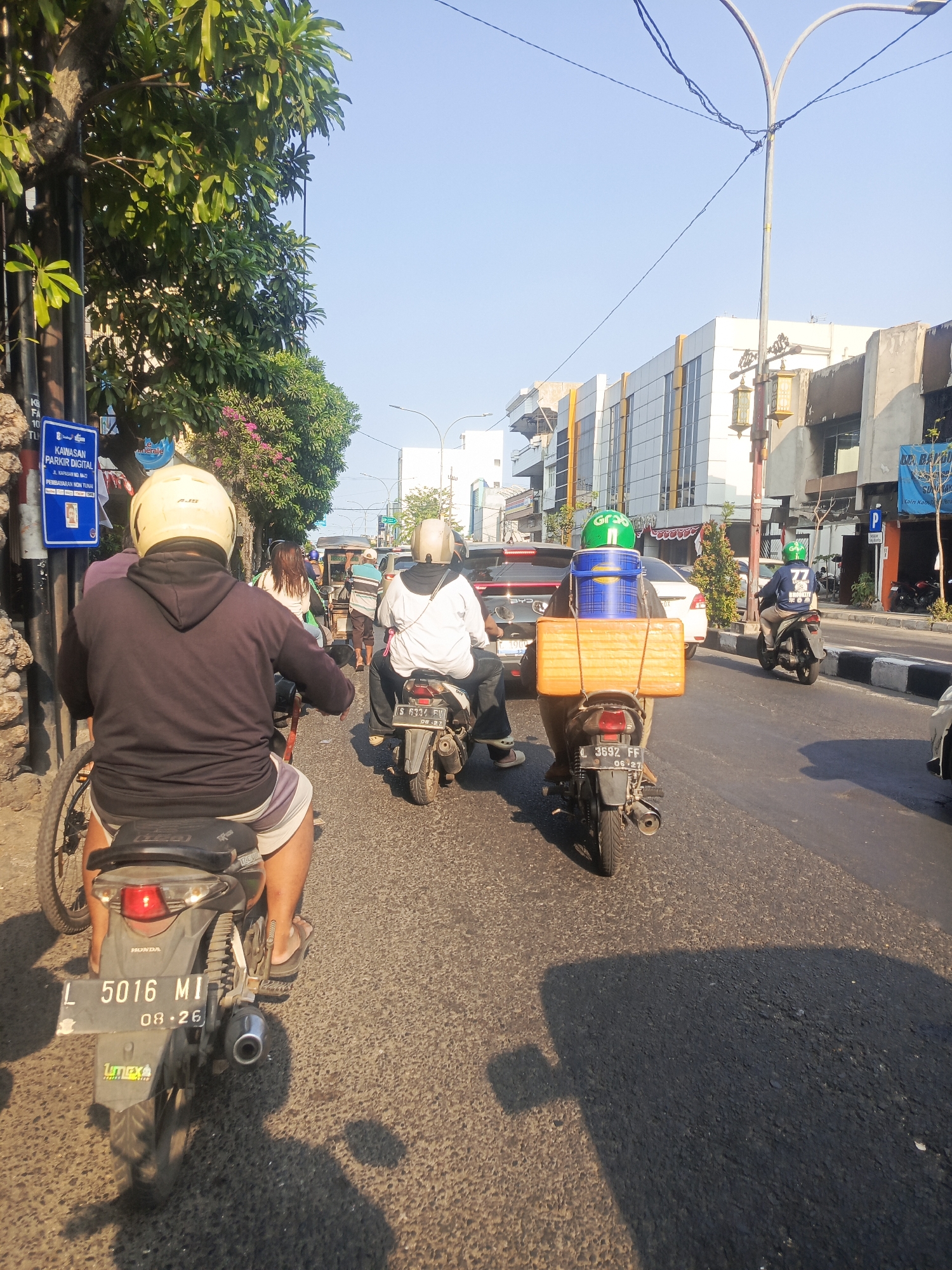 The image depicts a street scene in Surabaya, Indonesia, likely during the day, as indicated by the bright sunlight and clear sky. The street is moderately busy with traffic, primarily consisting of motorcycles. In the foreground, a person wearing a yellow helmet and a dark hoodie rides a motorcycle with the license plate "L 5016 MI". The motorcycle has a black seat and a visible exhaust pipe. The rider's legs are visible, wearing striped shorts and flip-flops.
Further down the street, another motorcycle is seen carrying a large orange box and a blue container on its rear rack, ridden by a person wearing a green helmet. To the right, a person in a white shirt and black pants, also wearing a helmet, is on a motorcycle. Other vehicles, including cars and more motorcycles, are present in the background, suggesting a typical urban traffic flow.
The environment includes a sidewalk with trees and vegetation, and buildings lining the street. A blue sign with text, possibly indicating parking regulations, is visible on the left side of the image. The road surface is asphalt, and shadows are cast by the vehicles and structures, indicating the direction of the sun. The overall impression is a snapshot of daily life and transportation in a bustling Indonesian city.