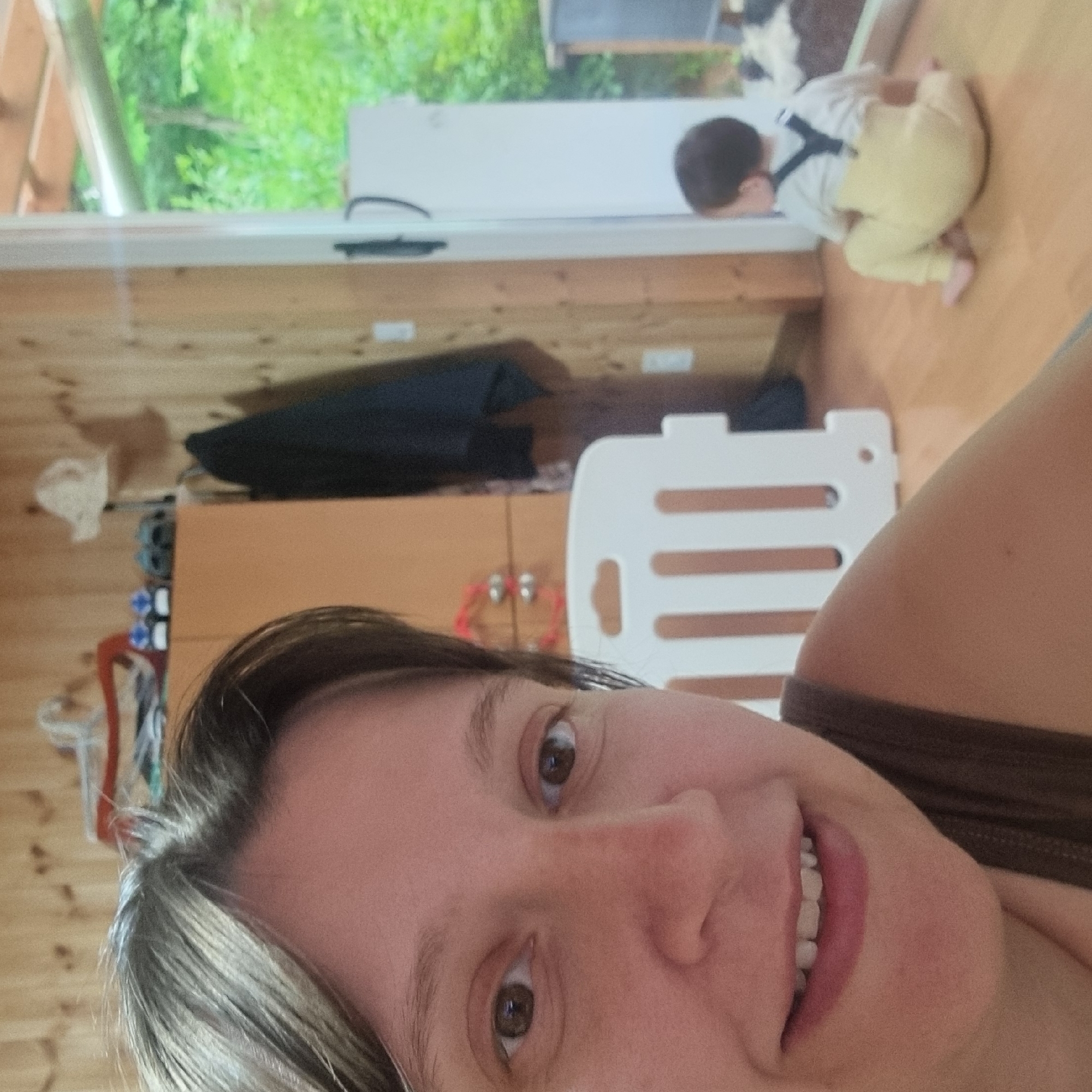 In an indoor setting with wood-paneled walls, a woman is taking a selfie. She has short, dark hair and is looking at the camera with a slight smile. In the background, a baby is sitting on the floor wearing a light-colored shirt and yellow pants with suspenders. A white playpen is visible between the woman and the baby. The room appears to be a living area or playroom, with wooden floors and a window showing greenery outside.