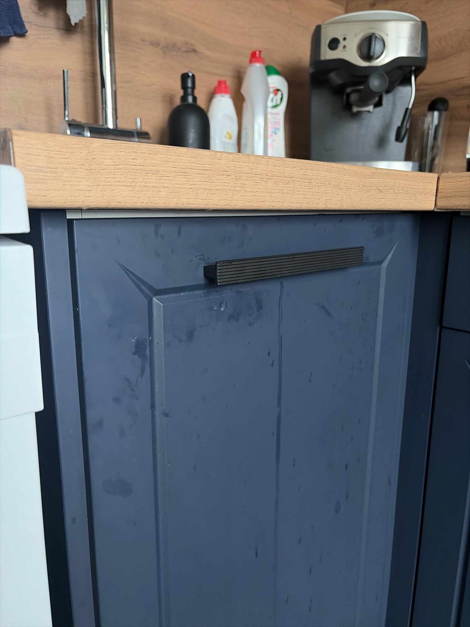 The image shows a close-up view of a dark blue kitchen cabinet. A black ribbed handle is attached horizontally to the cabinet door. Above the cabinet, a wooden countertop is visible, and behind it, a section of a wooden-paneled wall. On the wall, towards the right, a silver and black espresso machine is partially visible. To the left of the espresso machine, there are two bottles of cleaning product, one white and one red-capped. A faucet is also visible to the far left, mounted on the wall. The image does not contain any information about the city of Yekaterinburg, Russia.