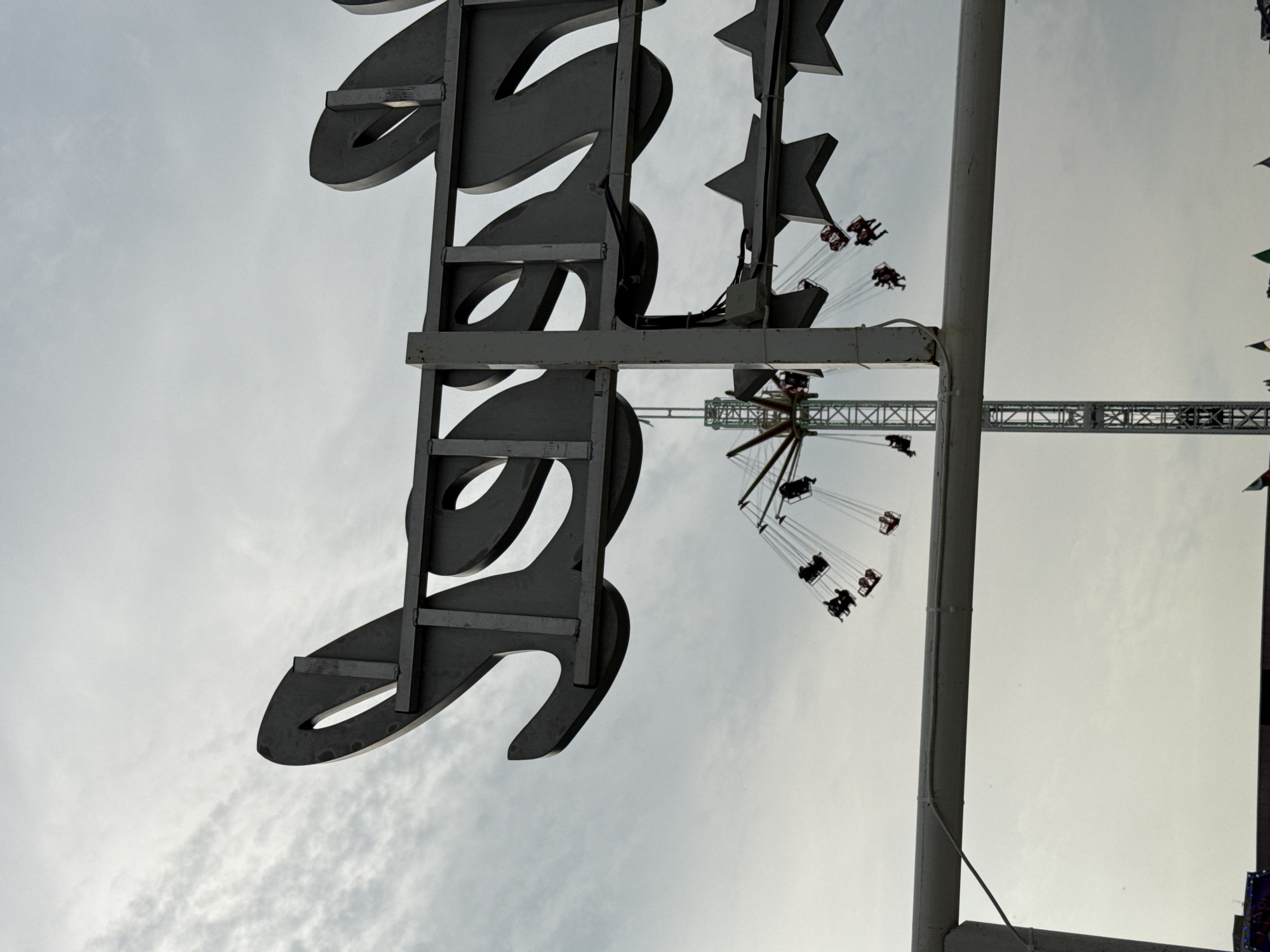 The image shows a low-angle shot of a fairground attraction, likely in Plovdiv, Bulgaria. In the foreground, a large, stylized metallic sign with stars is partially visible, obscuring the view. In the mid-ground, a swing carousel is rotating with people on the ride. A crane is visible in the background. The sky is overcast.