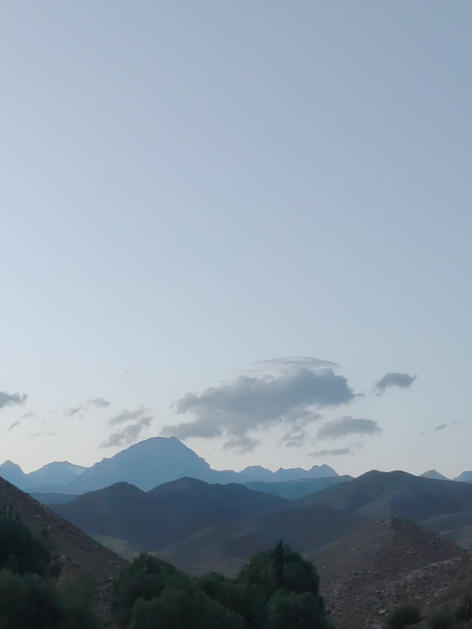 The image shows a landscape with several layers of mountains under a pale sky. The foreground features some greenery and rocky terrain. The middle ground consists of dark, undulating mountain ranges, and the background is dominated by a large, prominent mountain with a serrated peak. There are some wispy clouds in the sky. The overall scene depicts a natural mountain environment, likely in a rural area. The image does not contain any people or urban elements that would indicate the location is within the city of Cholpon-Ata, Kyrgyzstan.