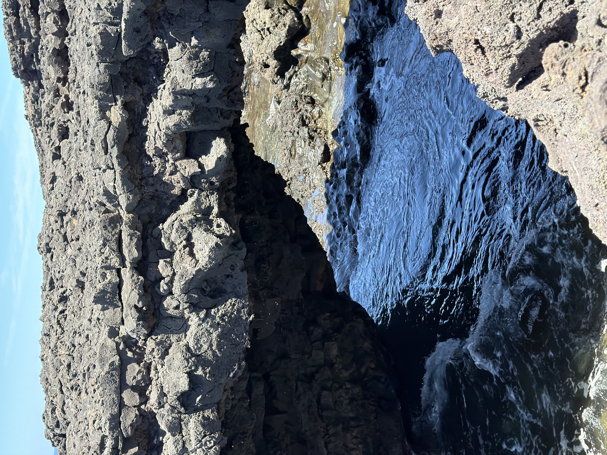 The image shows a narrow, water-filled crevice between two rocky formations. The rocks are dark and textured, with visible pores and cracks. The water in the crevice is dark blue with white foam, indicating movement and turbulence. The image appears to be taken at the Janubio Salt Flats in the city of Tinajo, Spain.