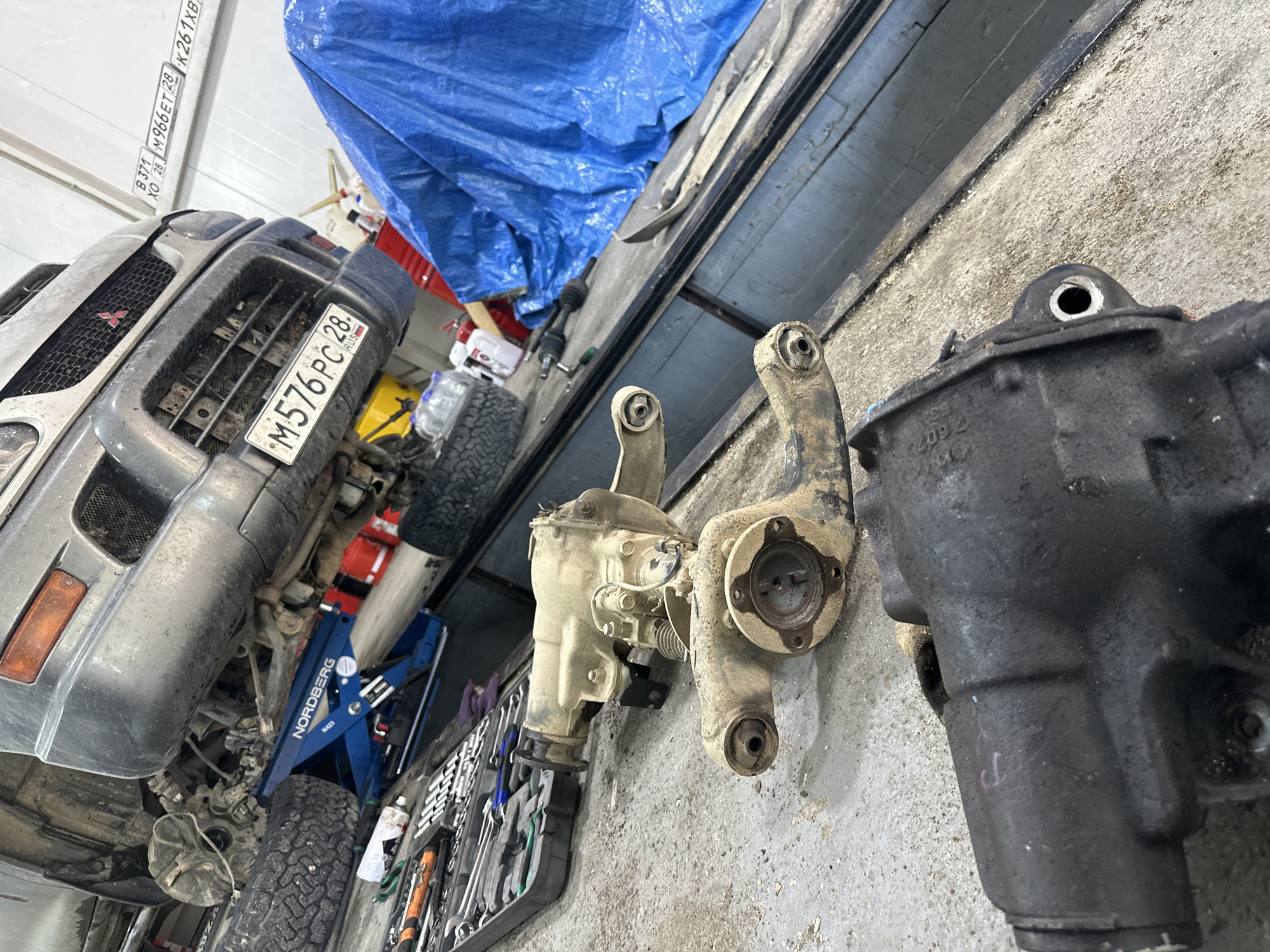 In a dimly lit auto repair shop, a grey Mitsubishi Pajero is elevated on a blue Nordberg hydraulic jack, its front end open for repair. Tools are scattered on the concrete floor, including a red toolbox and various wrenches. A spare tire is visible to the left of the vehicle. In the foreground, two differential assemblies are laid out on the ground: one is lighter in color and appears to be partially disassembled, while the other is a darker, possibly greasy, unit with red markings. A blue tarp covers an object in the background. License plates are visible on the wall, one reading "M576PC 28 RUSSIA". The overall scene suggests an ongoing automotive repair in Blagoveshchensk, Russia, given the Russian license plate and the typical appearance of a repair workshop.