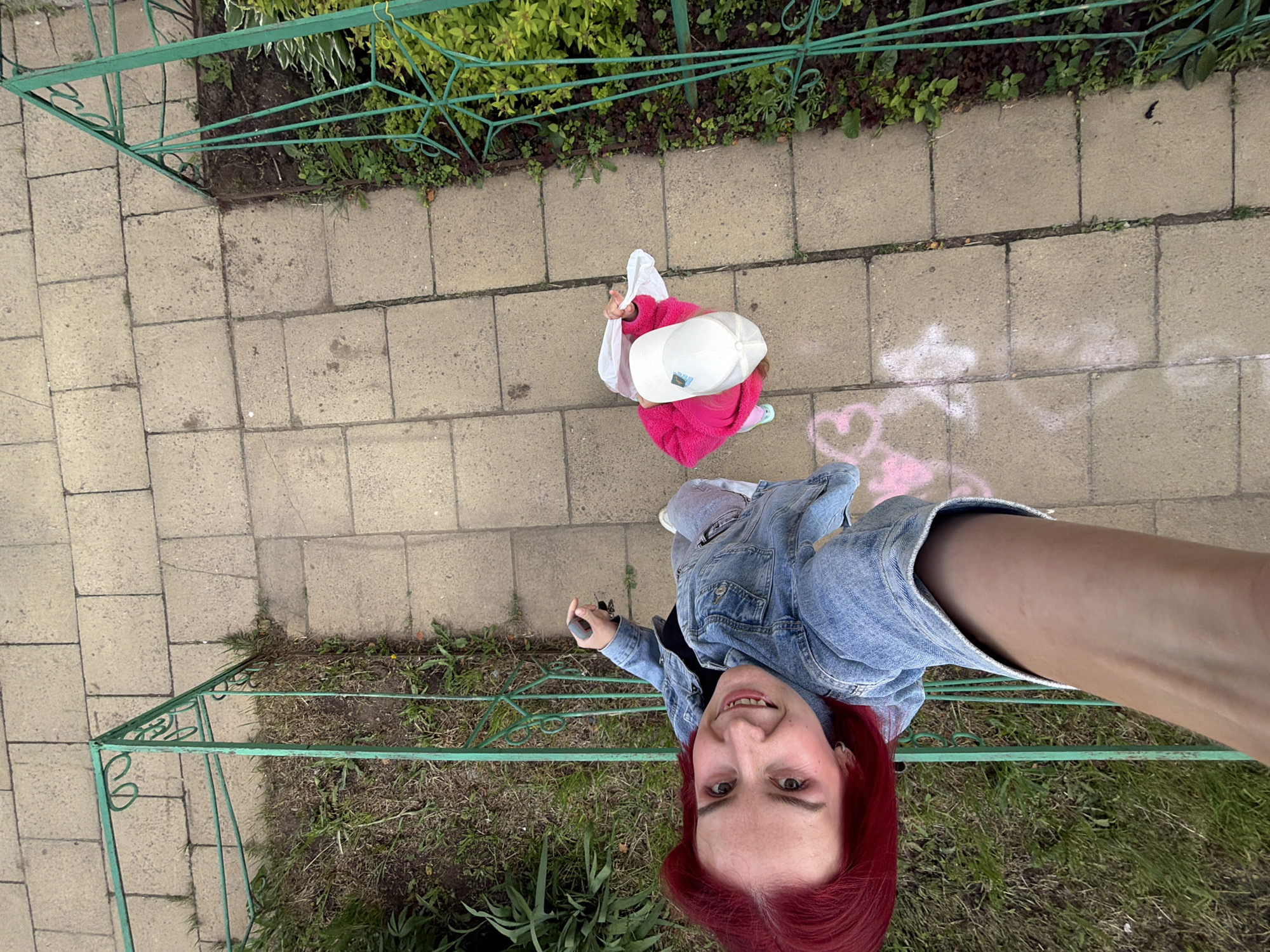 An overhead, slightly tilted selfie captures a woman and a child walking along a paved pathway in what appears to be a garden or park setting in Chaikovsky, Russia. The perspective is from above, looking down, with the woman's arm and face dominating the foreground.
The woman, with long red hair, wears a denim jacket and is smiling broadly at the camera. She holds a dark object, possibly a phone, in her hand. Ahead of her, walking away, is a child dressed in a bright pink fluffy top and a white baseball cap, holding a white plastic bag. The small logo or text on the child's cap is not clearly legible.
The pathway is made of rectangular grey paving stones. To the left and right of the pathway, there's a green metal fence or railing, partially obscured by lush green foliage and grass, indicating a garden or natural area alongside. Several pink and white chalk drawings, including a distinct pink heart, are visible on the pavement near the child and extending towards the woman's feet. The lighting suggests it is daytime, possibly overcast, providing soft illumination. The overall mood conveyed by the woman's smile is cheerful.
