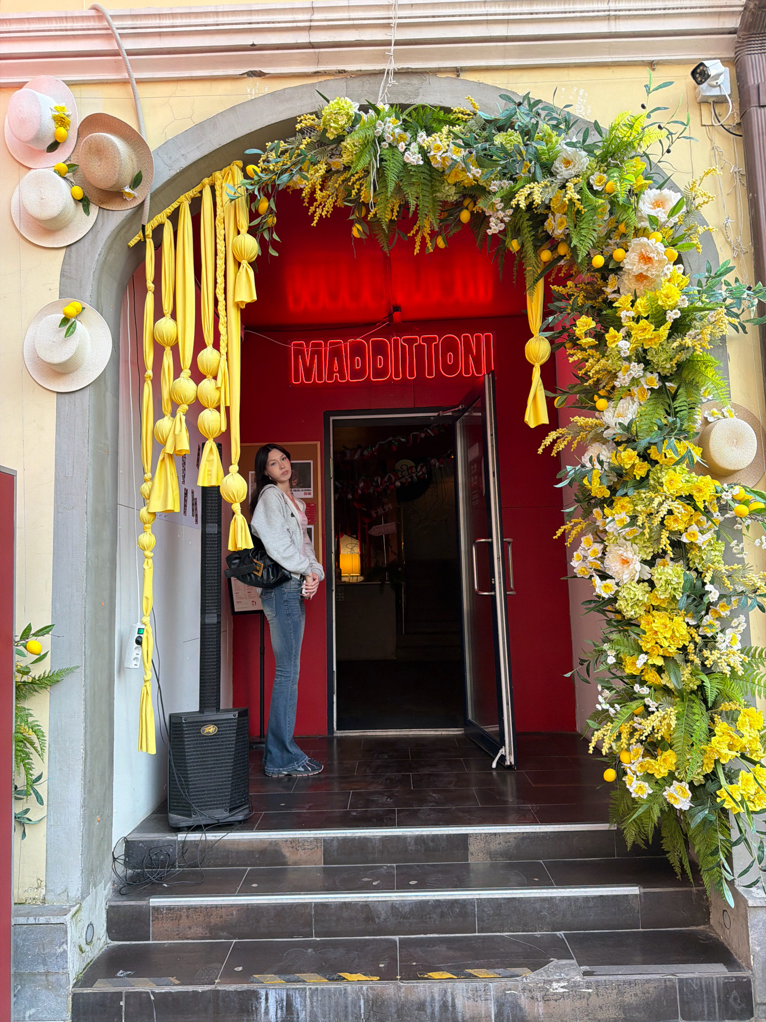 A young woman with dark hair, wearing a grey hoodie and blue jeans, stands on a tiled stairway in front of a red doorway. She is positioned to the left of the entrance, which is adorned with an archway of yellow and white flowers, greenery, and faux lemons. Above the doorway, a neon sign reads "MADDITTONI." To the left of the doorway, hanging yellow fabric streamers with knotted sections are displayed. Several straw hats with yellow flowers and lemons are affixed to the wall to the left of the entrance. A black speaker is placed on the stairway to the woman's left, with visible coiled cables. The overall scene appears to be an outdoor storefront decorated for a festive occasion. The location is identified as Moscow, Russia.