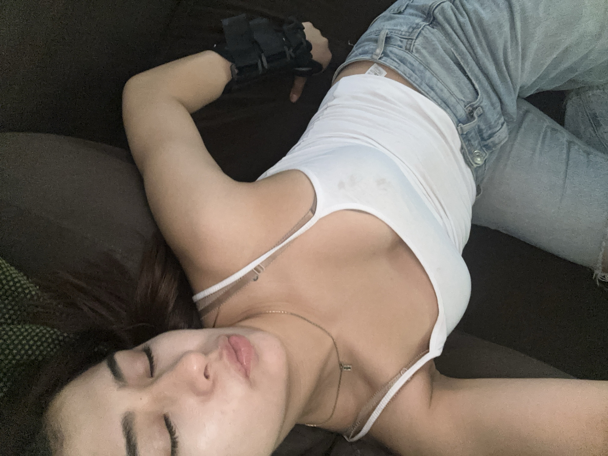 A close-up, slightly overhead indoor shot depicts a person, likely female, resting on a dark brown or black upholstered surface, such as a couch, in Puebla, Mexico. The individual has dark hair fanned out behind their head and light-to-medium skin. Their eyes are closed, and lips are slightly parted, suggesting a state of deep relaxation, sleep, or peaceful repose.
They are dressed in casual attire: a white spaghetti strap tank top and light blue ripped denim jeans. A delicate gold chain necklace with a small pendant adorns their neck. Notably, a black orthopedic brace or splint is worn on their right hand and wrist, indicating an injury or support need. The person's left arm is bent, with the hand out of frame.
Visible details include a white tag with unreadable black text, likely a brand or size label, peeking out from the waistband of the jeans. The background is indistinct, consisting only of the textured fabric of the furniture. The lighting is soft and consistent, characteristic of an indoor setting, and does not provide cues for the time of day or external weather conditions. The overall ambiance of the image is calm and serene.
