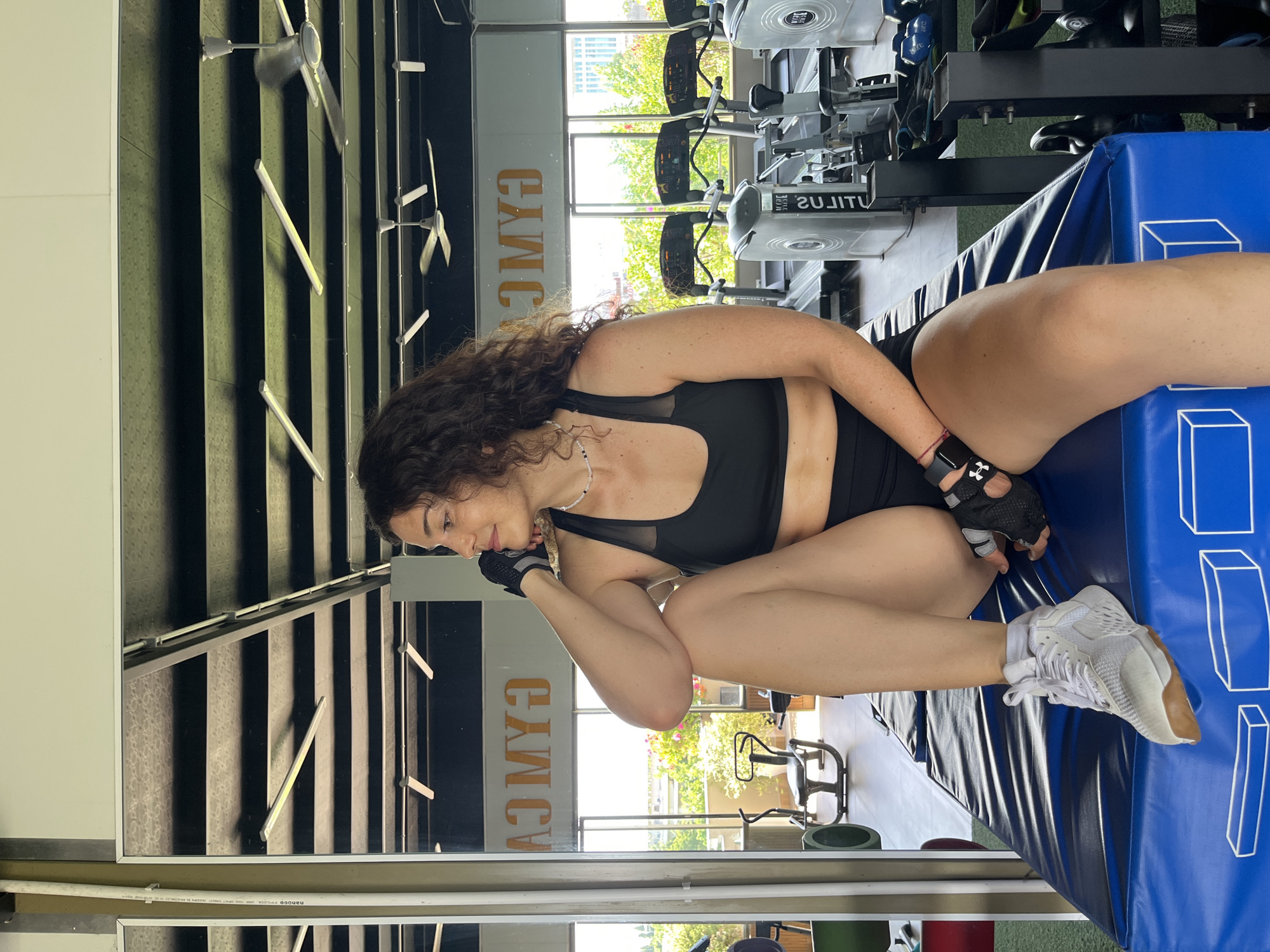 A woman with dark, curly hair is sitting on a blue mat in a gym. She is wearing a black sports bra, black shorts, white athletic shoes, and black workout gloves on both hands. Her left arm is bent, and she is resting her chin on her gloved hand, looking to her right. Her right leg is bent and positioned in front of her. Behind her, treadmills and weightlifting equipment are visible, as well as a large mirror reflecting the gym's ceiling with lights and ceiling fans, and the word "GYMC" written in orange letters. Through a large window in the background, trees and buildings can be seen, indicating an outdoor view. The scene is brightly lit, likely by natural light from the window. The gym appears to be located in Phan Thiết, Vietnam, as inferred from the presence of the word "GYMC" and the outdoor vegetation which is consistent with a tropical climate.