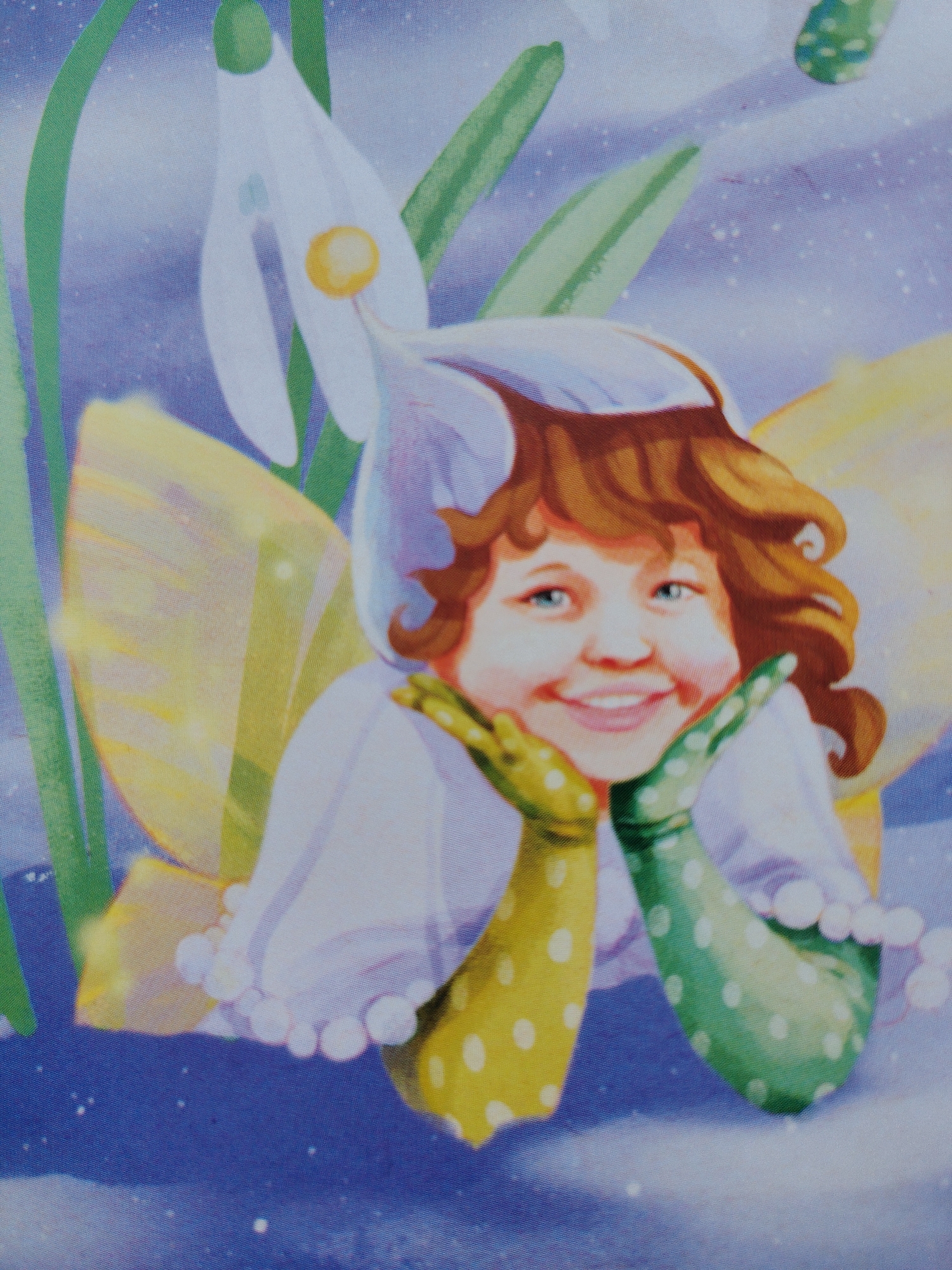 This is a close-up, digitally rendered illustration of a young fairy with curly brown hair, blue eyes, and a broad smile. She is wearing a white outfit with a ruffled collar and has yellow and green spotted sleeves. Her hands are resting on her chin, and she appears to have translucent, yellow wings. She is surrounded by stylized snowdrop flowers with green leaves against a light blue, speckled background that suggests a wintery or magical setting.