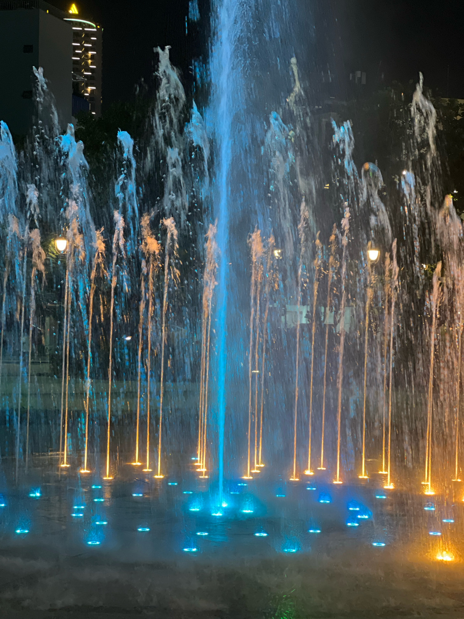 The image shows a night view of a fountain in Nha Trang, Vietnam. The fountain features multiple streams of water illuminated by vibrant blue and orange lights, creating a dynamic and colorful display. Some lights are submerged in the water, while others are positioned at the base of the water jets. In the background, a tall building with illuminated windows and a prominent yellow logo can be seen, along with trees and streetlights, indicating an urban setting. The overall scene is one of nighttime entertainment and urban ambiance, likely a public space designed for aesthetic appeal and visitor engagement.