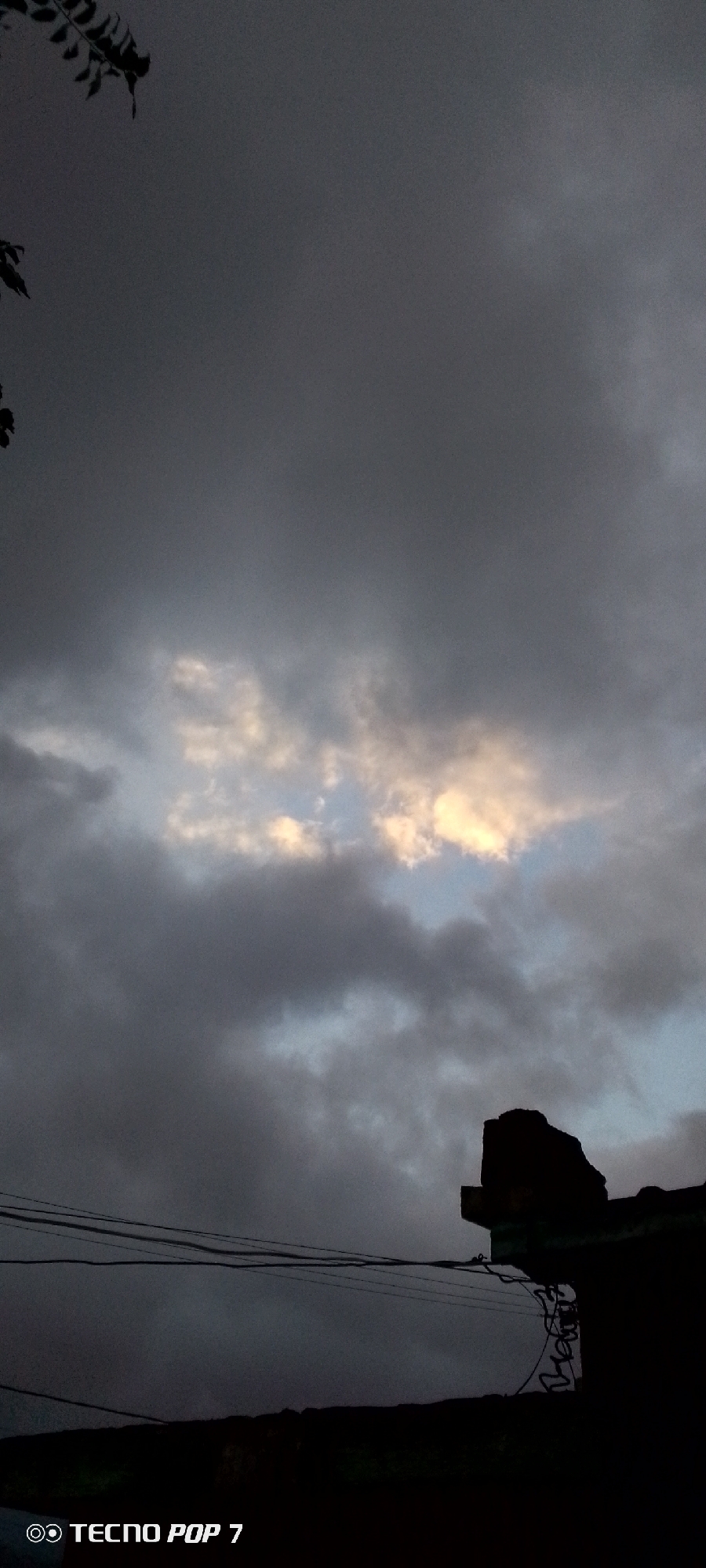 The image displays an overcast sky, filled predominantly with heavy, dark grey clouds. Towards the center-right, a patch of lighter, yellowish-white clouds stands out, suggesting indirect sunlight breaking through or reflecting off them, with a small area of pale blue sky visible in between.
In the top-left corner, dark, silhouetted foliage from an unseen tree or bush extends into the frame. The lower portion of the image reveals man-made structures: several dark power lines or communication cables stretch horizontally across, leading towards a dark, indistinct silhouette of a building or structure in the bottom-right. Coiled wires are also visible near this structure.
The overall dim lighting points to either an early morning, late afternoon, or a heavily overcast day, hinting at inclement weather like impending rain. The scene evokes a somewhat solemn or anticipatory mood due to the dense cloud cover.
A watermark in the bottom-left corner clearly reads "◎ TECNO POP 7," identifying the camera device. The presence of power lines and buildings is consistent with an urban or semi-urban setting, fitting the provided location context of Altagracia, Venezuela. No specific activity or people are visible.