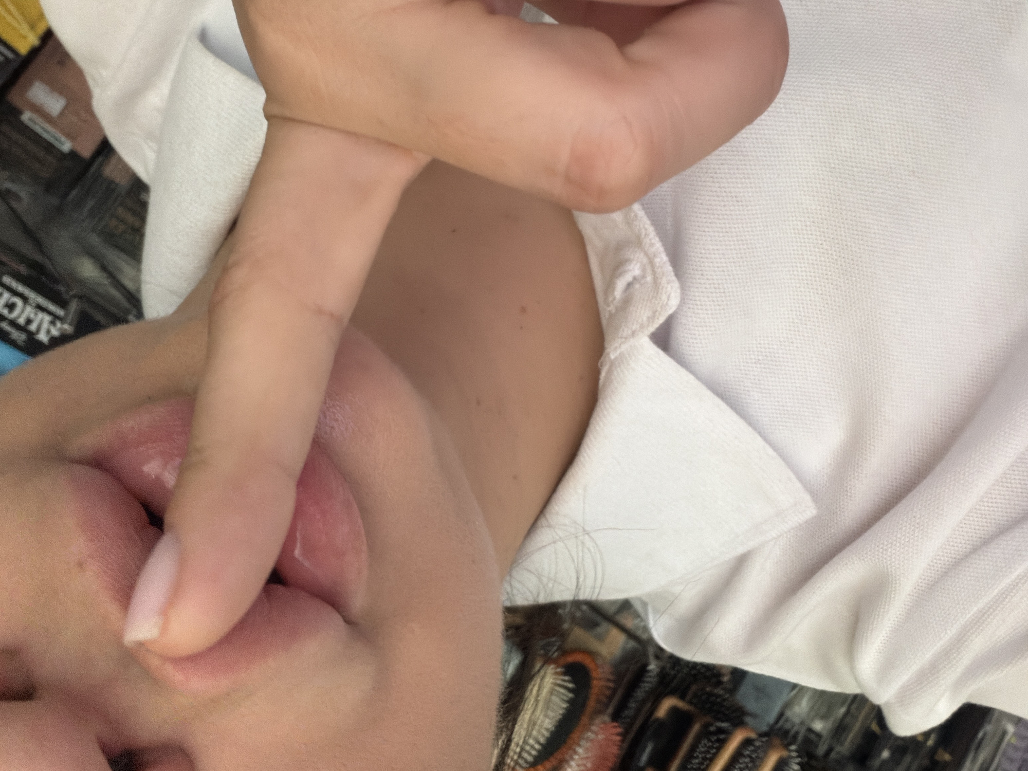 A close-up, vertically oriented image depicts a person's fair-skinned lower face, including the chin and lower lip, along with a hand. A finger and thumb are actively pressing against and lightly pinching the lower lip and chin area, causing a slight indentation and distortion of the lip. The fingernail on the visible finger is pale and appears neatly manicured. Several small moles or freckles are visible on the skin of the neck and chin.
The person is wearing a white collared shirt, likely a polo shirt, distinguished by its textured, pique-knit fabric, which is visible on the right side of the frame and under the chin. Strands of dark hair are present near the collar and along the jawline.
The background is blurred, indicating an indoor setting. Dark, indistinct objects, possibly merchandise or packaging, fill the out-of-focus areas. Partial white text, including the word "ALICE," is discernible on one of these dark background elements.
The gesture of touching the lip and chin could suggest contemplation, nervousness, or a habitual action. While the location context is given as Sochi, Russia, no specific visual cues within the image confirm this geographical setting.