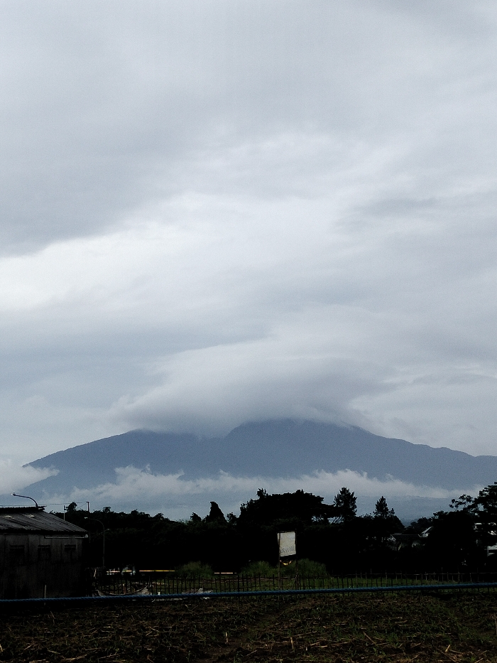 The image displays an overcast and moody landscape in Dramaga, Indonesia. A large mountain or hill dominates the background, partially enveloped by thick, low-hanging clouds that blend into the uniformly gray sky. The mountain appears in dark blue-gray tones, with its contours softened by the cloud cover.
In the mid-ground, a dark, silhouetted band of trees and dense foliage stretches across the frame. To the far left, a dark, weathered building, possibly a shed or a small structure with a corrugated roof, is visible. Amidst the trees on the right side, a light-colored, rectangular sign stands out, though any text on it is too small and blurred to be legible. A horizontal structure, resembling a low fence or barrier, runs across the mid-ground, with a dark blue pipe or hose visible beneath it.
The foreground consists of a dark, possibly uncultivated or barren field with visible texture on the ground. The overall lighting is dim, indicative of a heavily overcast day, contributing to a quiet, somber, and somewhat mysterious atmosphere. There are no people or discernible activities taking place. The primary colors in the scene are various shades of gray, dark blue, and black.