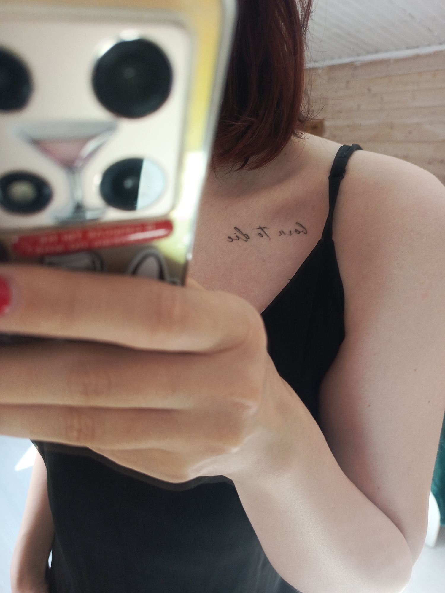 A person is holding a cell phone in front of them, partially obscuring their face and body. The phone has a decorative case featuring a martini glass and black circles. The person has short red hair and is wearing a black tank top. A tattoo is visible on the upper chest near the collarbone. The background appears to be a room with light-colored wood paneling.