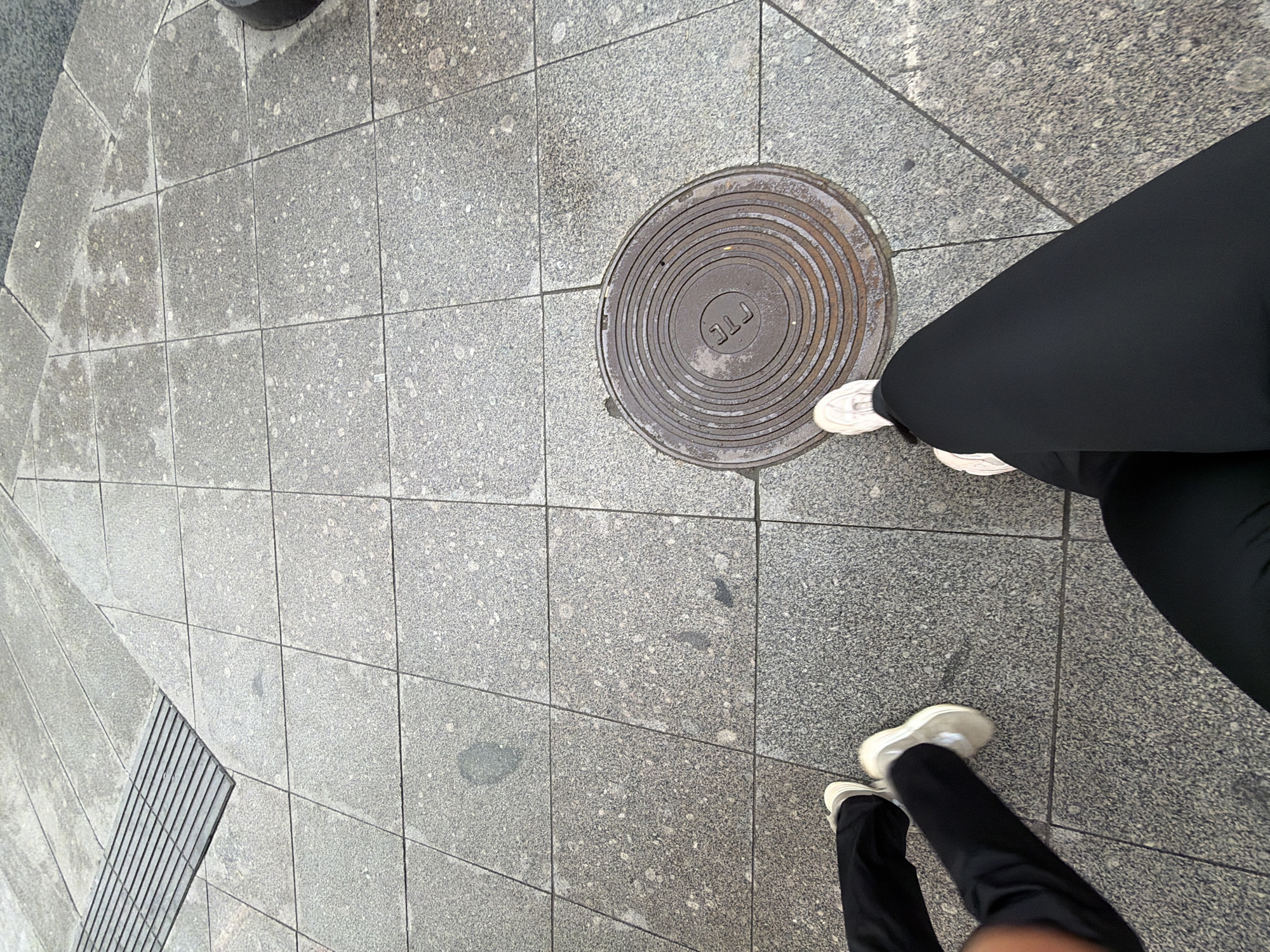 A high-angle shot depicts a pedestrian walking on a paved urban surface. The surface comprises square, gray-toned concrete tiles. A circular manhole cover is embedded in the pavement. The pedestrian is wearing dark-colored athletic leggings and white sneakers. The scene suggests a city street or sidewalk environment. The location in Saint Petersburg, Russia, cannot be verified with the given image.