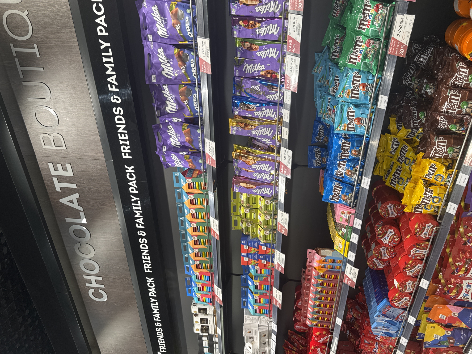 The image shows a shelf in a chocolate boutique, displaying various chocolate products, with the label "CHOCOLATE BOUTIQUE FRIENDS & FAMILY PACK" visible on the side. Several chocolate brands are visible, including Milka and M&M's. The chocolate bars and packs are arranged on the shelves. Price tags and "save" percentages in RUB (Russian Rubles) are visible for some items. The variety of chocolates includes assorted flavors and types. The shelf appears to be located in a store in Sochi, Russia, based on the presence of Russian currency (RUB) and chocolate brands commonly found in the region.