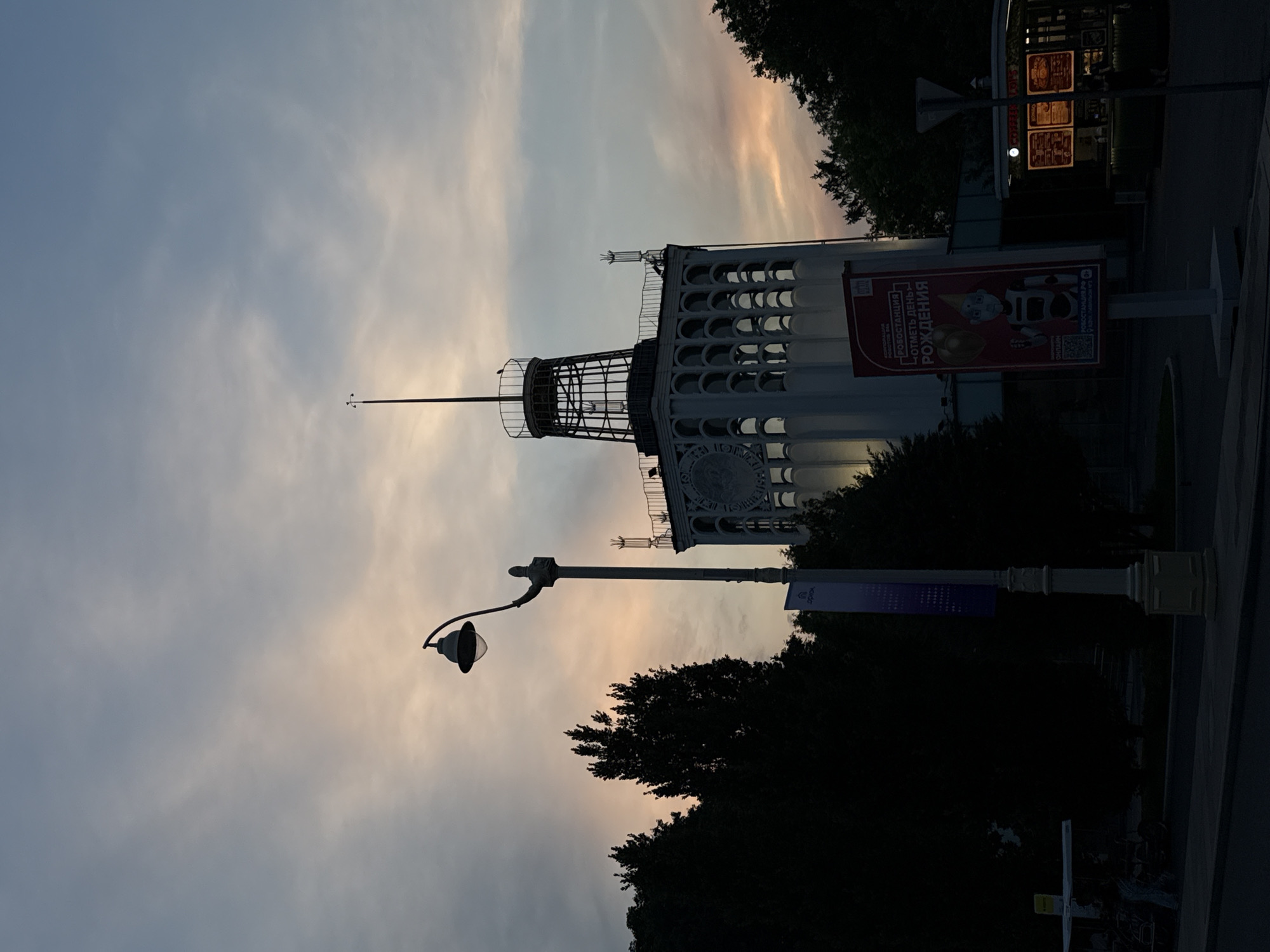 The image depicts a dusk scene in Moscow, Russia, featuring the iconic Pavilion No. 66 "CULTURE" at VDNKh. A streetlamp stands in the foreground, its silhouette stark against the pastel-hued sky. Behind it, the distinctive tower of Pavilion No. 66 rises, characterized by its circular windows and decorative spire. Dark silhouettes of trees frame the scene, contributing to the twilight atmosphere. A billboard advertising "ROBOSTATION" and a small kiosk with illuminated menus are visible to the right of the pavilion. The sky exhibits soft clouds tinged with orange and pink, indicative of a sunset.