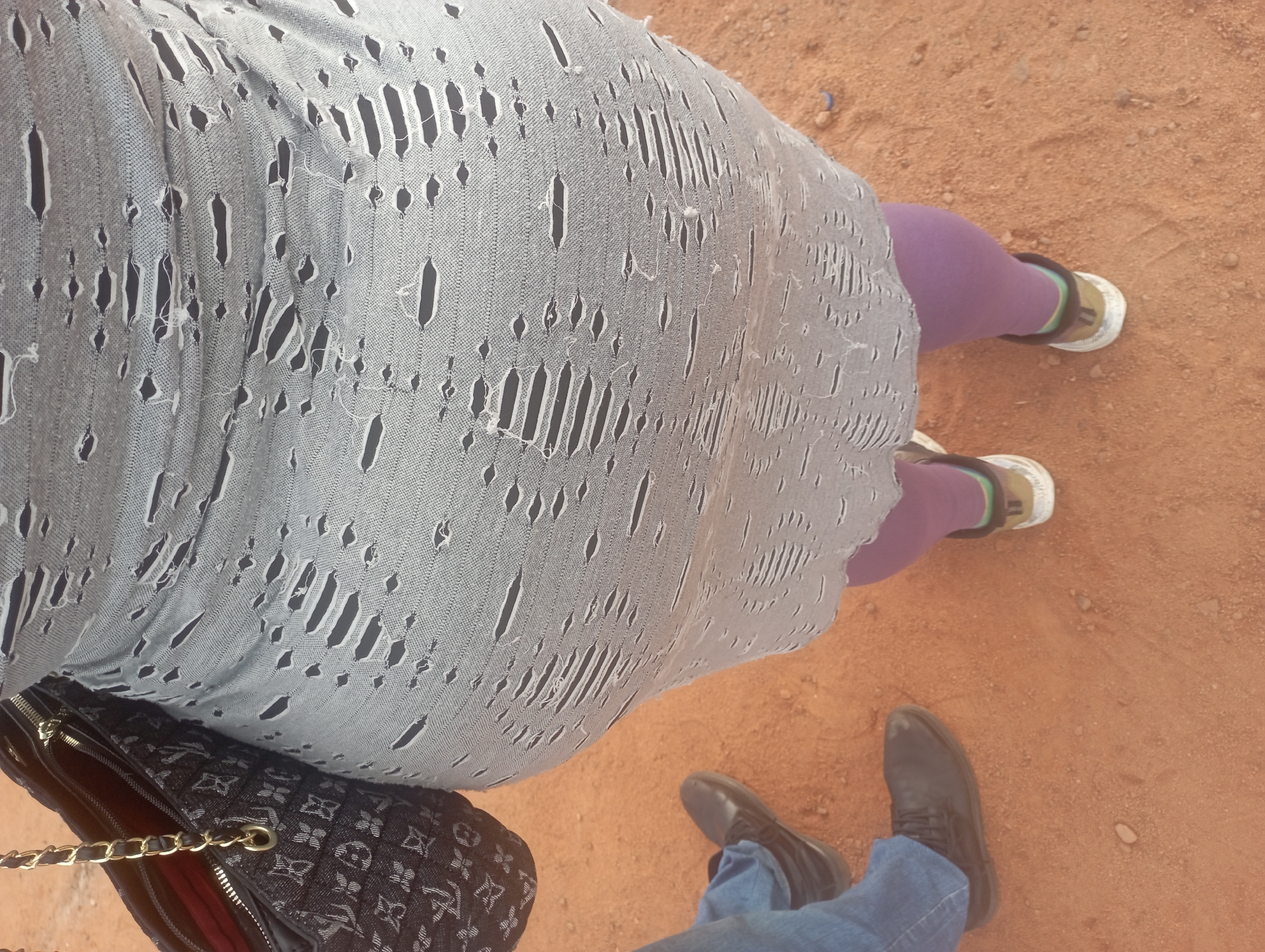 The image shows a close-up of two people standing on a dirt ground, likely outdoors. One person is wearing a grey, distressed garment with numerous vertical slashes, paired with purple leggings and athletic shoes. The other person is wearing blue jeans and dark dress shoes. A black purse with a gold chain strap and a repeating monogram pattern is visible on the left side of the frame, resting on the ground. The ground appears to be dry, reddish-brown dirt with some small pebbles and indentations, possibly footprints. The lighting suggests it might be daytime, with no clear indication of weather conditions. The focus is on the lower half of the individuals and their attire, creating a ground-level perspective.