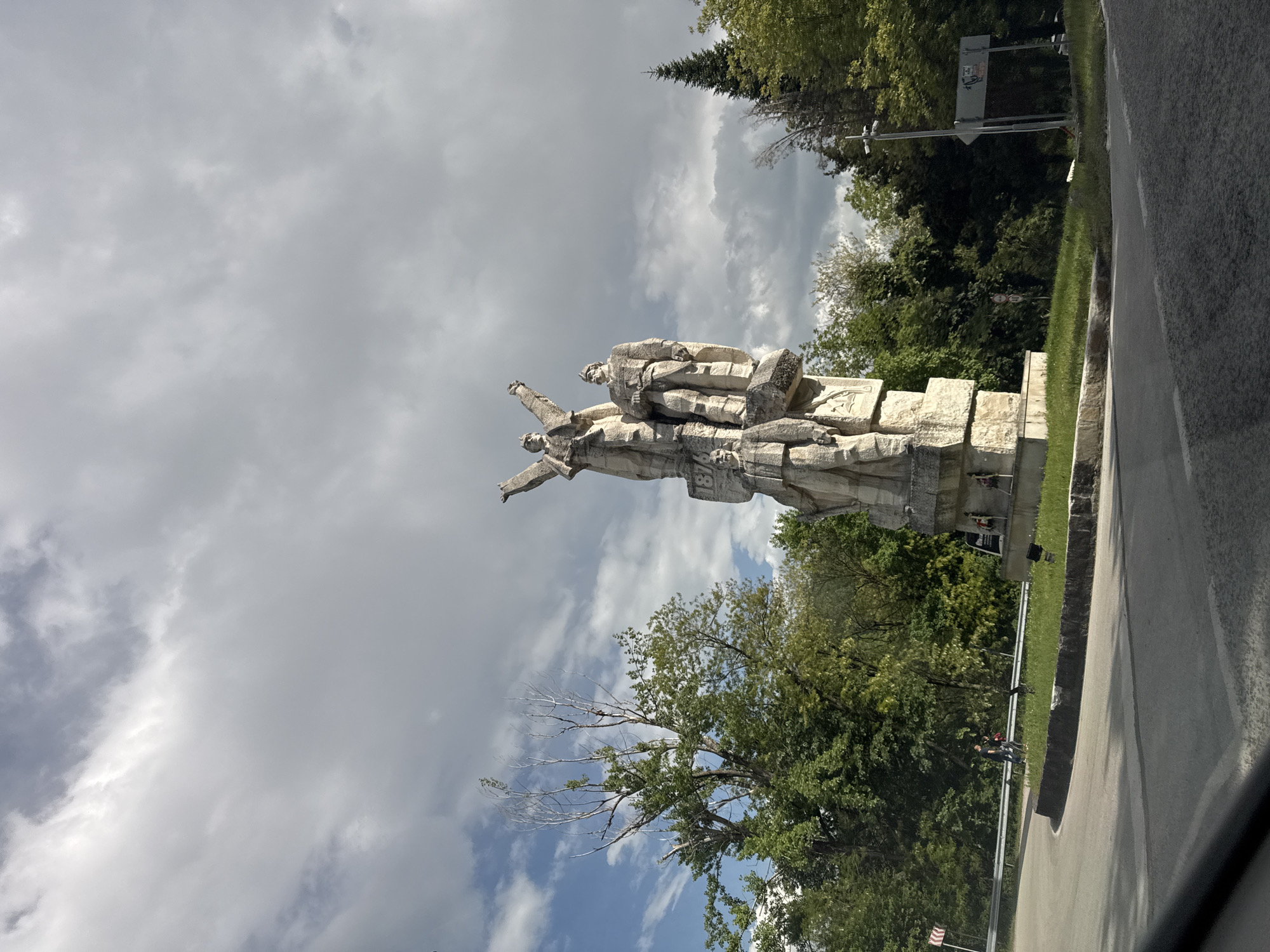 The image shows a large stone monument in Gesha, Bulgaria. The monument depicts three figures in dynamic poses. It is situated on a stone platform next to a road, surrounded by trees and grass. The sky is cloudy. Two people can be seen walking near the monument. A road sign is visible in the background.