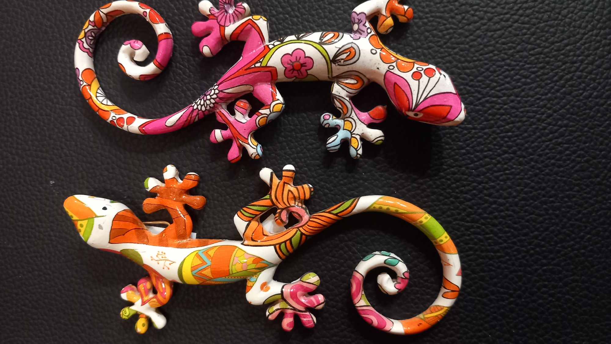 Two decorative ceramic geckos are positioned on a textured black surface. The gecko on the left is primarily orange, white, and green, with intricate patterns. The gecko on the right features pink, white, and floral designs. Both geckos have curled tails and spread-out limbs. The lighting is such that shadows are cast on the surface, indicating the direction of the light source. The overall scene is that of an indoor display, likely a retail environment or a home decoration. There is no indication of a specific location within the city of Kos, Greece, in the provided image or its crops.