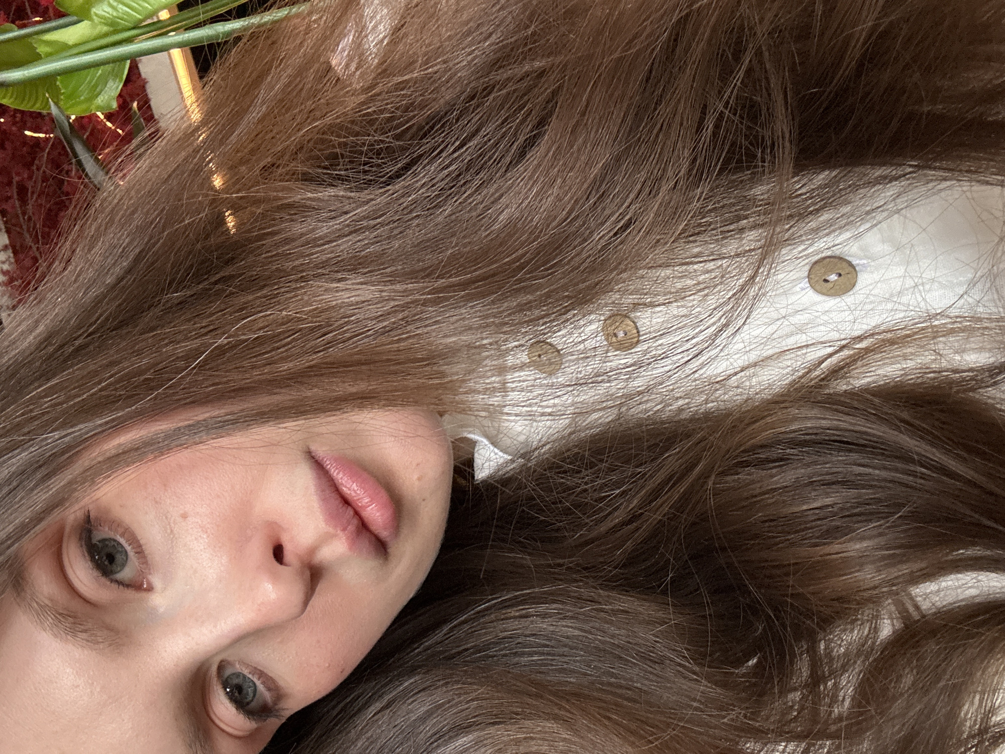 A close-up, angled selfie features a woman with brown hair, partially obscuring her face. Her eyes are visible, and she has fair skin and pink lips. She is wearing a white garment with brown buttons. A plant with green leaves is visible in the top left corner. The setting is ambiguous. No specific location can be determined.
