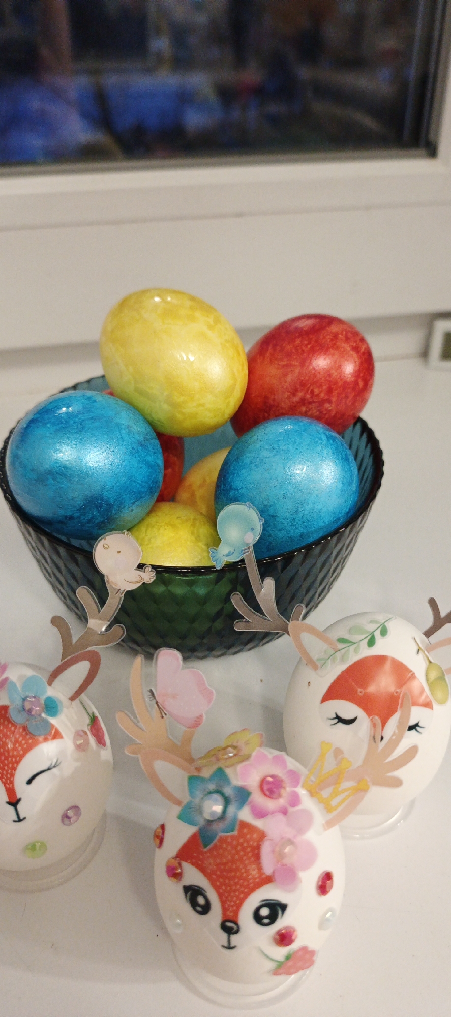 The image shows a still life of Easter eggs. A dark green textured glass bowl is filled with colored Easter eggs in shades of blue, yellow, and red. In front of the bowl are three decorated Easter eggs, each shaped like a deer with painted faces, antlers, and embellishments like flowers, jewels, and a butterfly. The background includes a window, which likely means this is a photo taken indoors in a house.
The information about the city of Chaikovsky, Russia is not evident in the image.