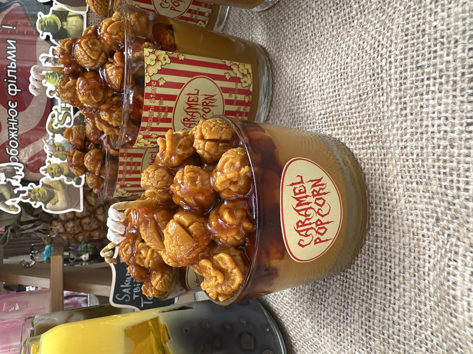 The image displays a close-up view of several glass containers filled with decorative items designed to resemble caramel popcorn, positioned on a light brown woven fabric surface. In the foreground, a prominent glass container features a light brown base topped with a darker, glossy caramel-like substance covering numerous textured, brownish-orange spheres that mimic popped corn kernels. A gold-rimmed oval label with red, stylized text reading "CARAMEL POPCORN" is affixed to the front of this container. Directly behind it, slightly to the right, another similar glass container is visible, featuring a red and white striped label with text including "Shimauchi Store," "«light in the dark»," "CARAMEL CORN," and a partially visible "50 hour burn," indicating these are likely scented candles. Additional similar containers are partially visible further back. To the far left, a clear glass bottle contains a layered liquid, bright yellow on top and a darker, possibly black liquid at the bottom, with small, dark circular objects suspended within the darker layer. The background includes a partial view of a small black chalkboard sign with white handwritten text reading "Sakura TBI T," and a display featuring cardboard cutouts of characters from the Shrek animated film franchise. Above the characters, a banner with Cyrillic text, including "обожнює фільми !" (translating to "loves films!"), is visible. A light wooden stand and a large dried pineapple-like object are also partially discernible in the background. No individuals are present in the scene, which appears to be a retail or display setting in Unknown, Unknown.