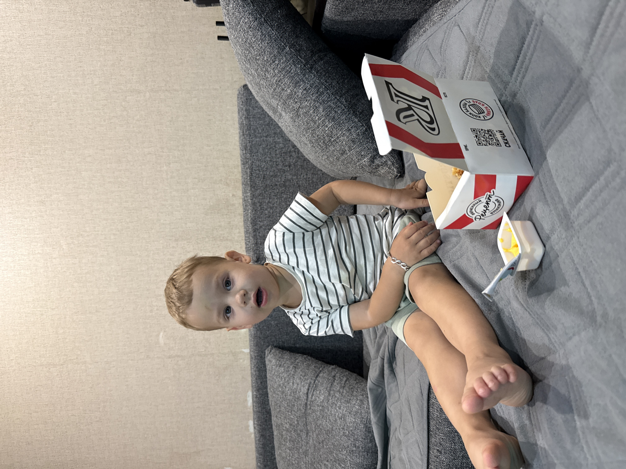 In a dimly lit room, a young boy with blond hair and blue eyes sits cross-legged on a gray sofa. He is wearing a striped t-shirt and light-colored shorts. In front of him, to his right, sits a white and red fast-food container with food inside. A small plastic container with yellow sauce and a spoon is placed to his left. The background consists of a textured beige wall and a darker gray pillow on the sofa. The overall scene suggests a casual setting, possibly a living room, where the boy is enjoying a meal. The location, Rusanovo, Russia, is not visually evident from the image itself but is provided as contextual information.