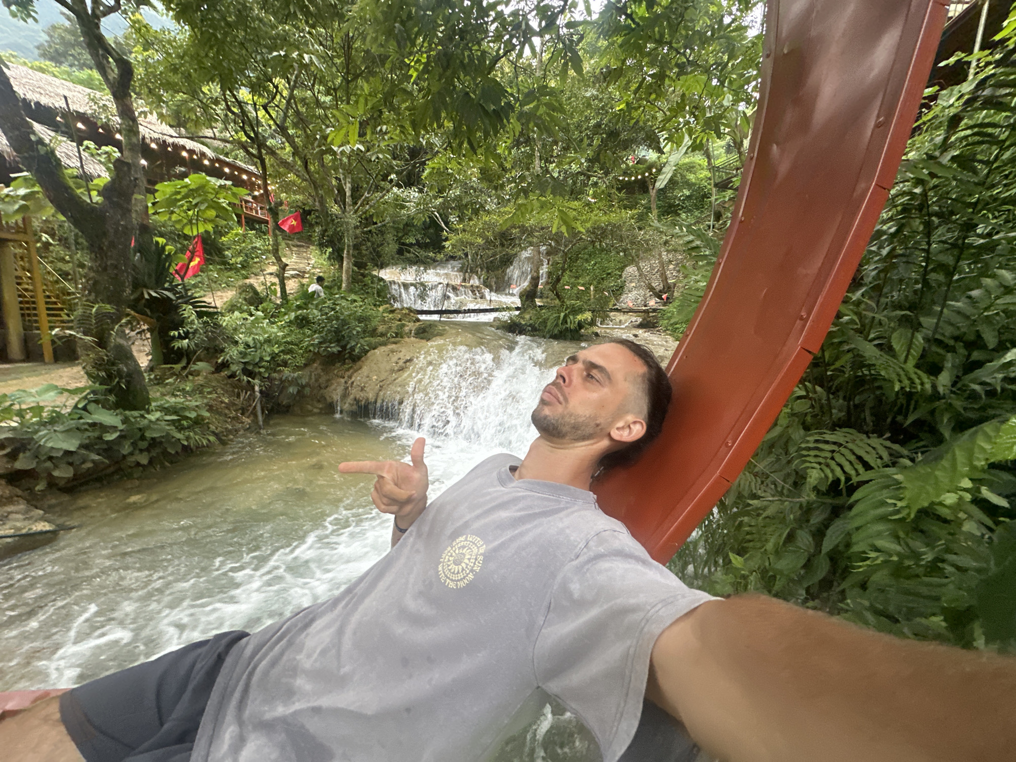A man lies on a red slide next to a waterfall. He is wearing a grey t-shirt and dark shorts. The t-shirt has a circular graphic with text that reads "MADE WITH THE SUN IN THE MIDDLE OF THE SUMMER". The man is looking up and to his left, with his right arm extended, pointing towards the waterfall. The waterfall cascades over rocks into a shallow river. Lush green foliage surrounds the scene, with trees and ferns creating a natural, tropical environment. In the background, there are glimpses of a traditional building with a thatched roof and decorative lights, along with several red flags. The scene appears to be outdoors during the daytime, with the lighting suggesting it could be overcast or shaded. The overall impression is one of leisure and enjoyment in a scenic, natural setting. The man's pose and expression suggest a relaxed or playful mood.