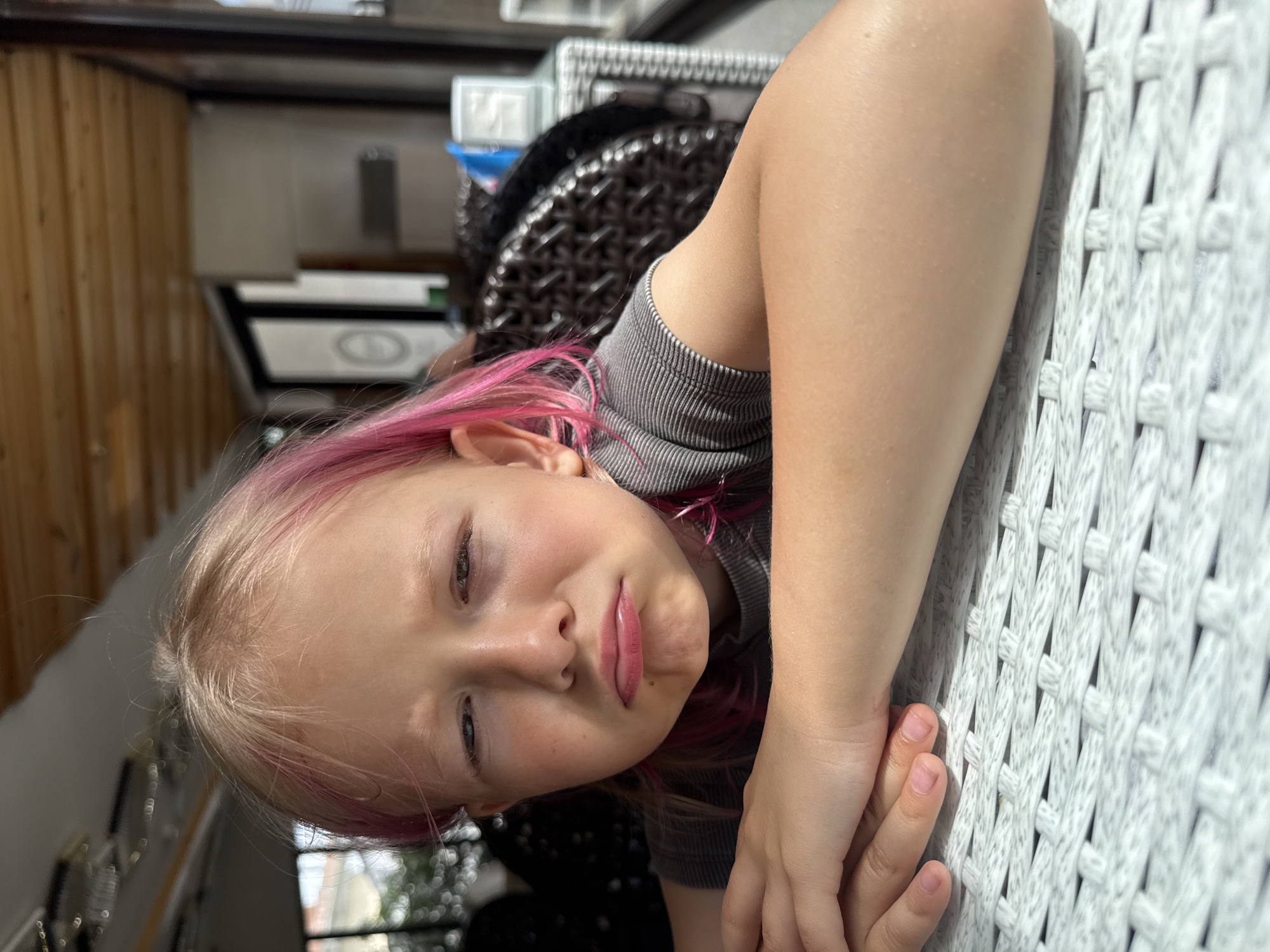 A young girl with light skin and blonde hair, accented with significant pink streaks, lies down and looks towards the camera. She has a relaxed expression with a subtle pout on her lips and her eyes appear a light color. She is wearing a grey ribbed short-sleeved top. Her head is tilted, giving the impression she is lying on her side, and her left arm rests upon a prominent white, intricately woven surface, likely a chair, bench, or sofa. This white woven pattern occupies a large portion of the right side of the image. In the blurred background to the left, wooden wall paneling is visible. Behind her head and shoulder, a dark, textured or patterned object, possibly another piece of furniture, can be seen. The scene appears to be set indoors or in a covered outdoor area during the daytime, suggested by the bright, natural light. No text is visible.