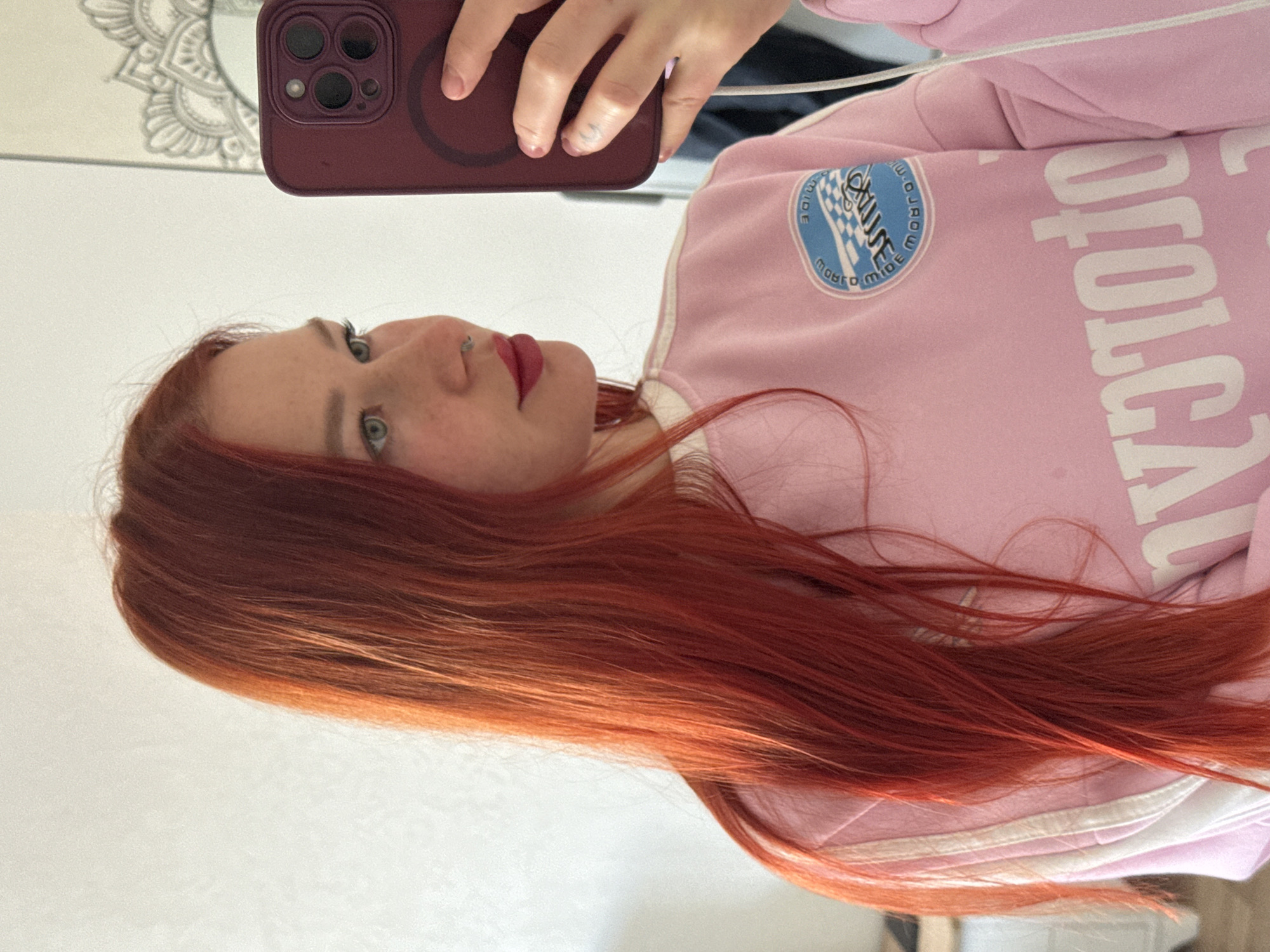 A close-up mirror selfie captures a person, likely female, with long, flowing bright red/auburn hair. Her head is tilted, showing her left profile, with striking light-colored eyes (appearing green/blue) and dark red lipstick. She has a septum piercing.
The person is wearing a light pink sweatshirt or jacket, featuring white stripes along the arm and side. On the left chest area of the garment, there is a circular blue and white emblem. This emblem clearly displays the word "CRUISE" above a checkered flag, and below it, the word "WORLDWIDE" is partially visible, oriented upside down in the reflection. Large white letters are visible on the main body of the sweatshirt, but they are mirrored and partially obscured, making them difficult to decipher fully. When read directly from the image, they appear as "OYOTOD" or similar.
She is holding a dark reddish-purple iPhone in her hand, visible in the upper part of the frame, indicating the selfie is taken in a mirror. The phone has multiple camera lenses and a visible MagSafe ring on its back.
The background is predominantly a plain white or light-colored surface, likely a wall or another mirror. A decorative mandala-like pattern is reflected in the upper left corner, behind the person's head. The entire image is rotated 90 degrees clockwise. The setting appears to be indoors.
