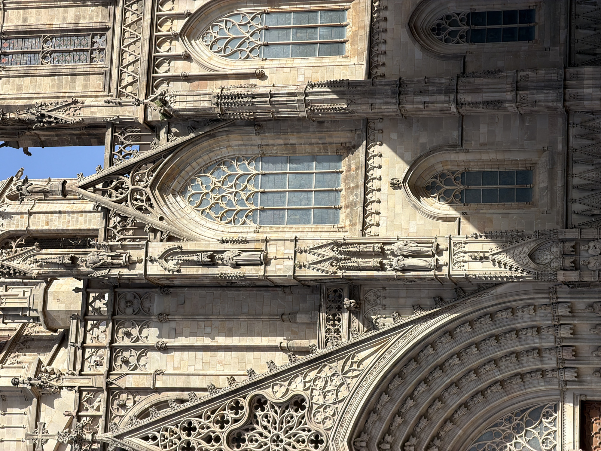 The image captures a section of the Barcelona Cathedral, also known as the Cathedral of the Holy Cross and Saint Eulalia, in Barcelona, Spain. The façade is constructed of weathered stone blocks, showcasing Gothic architectural elements. Ornate carvings, including statues of figures in robes and what appear to be gargoyles, adorn the building. Large, arched windows with intricate tracery are visible, some filled with stained glass. The sunlight casts strong shadows across the textured stone surfaces, highlighting the depth and detail of the architecture. The sky above is a clear blue.