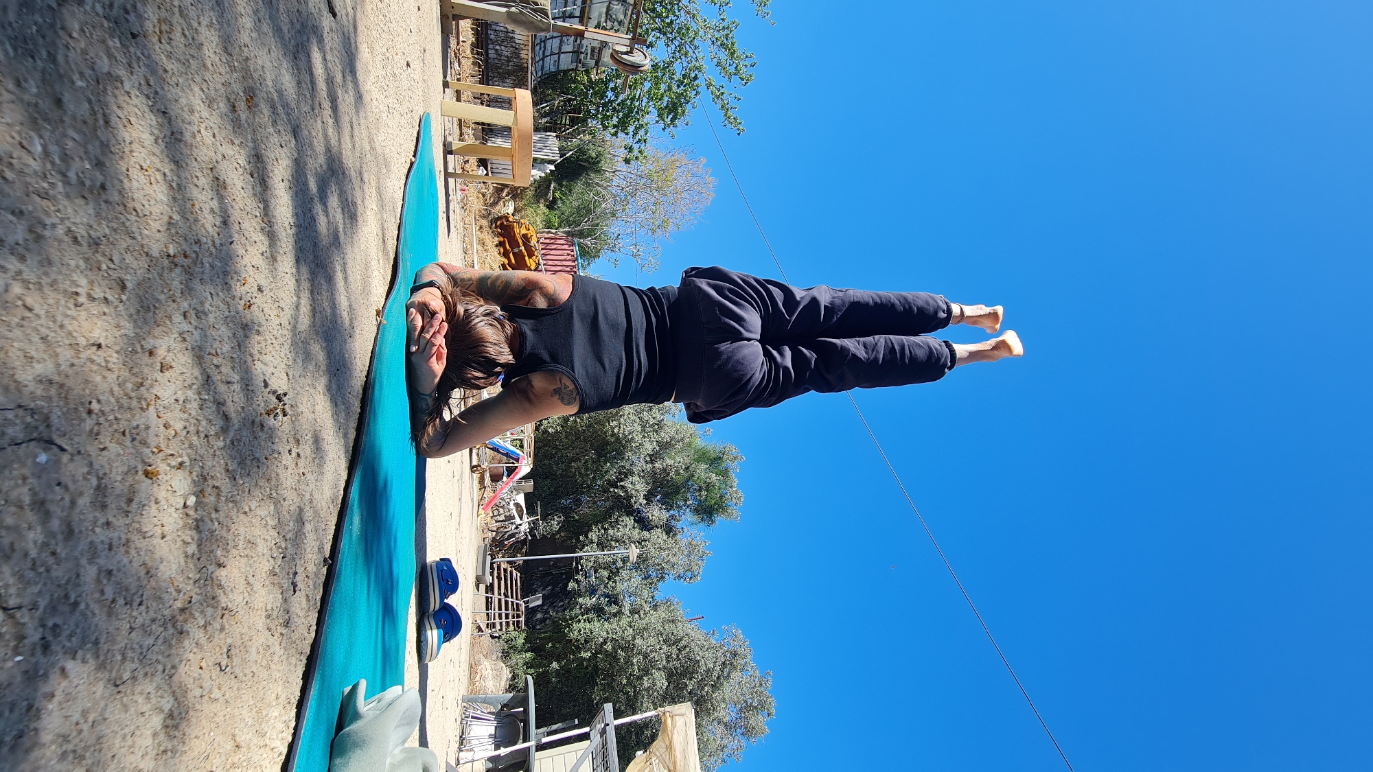 An outdoor, eye-level shot captures a person performing a yoga or fitness pose under a clear blue sky. The individual, seen from behind, has medium-length brown hair and is wearing a black tank top and dark sweatpants. They appear to have tattoos on their upper back and shoulders. They are lying prone on a bright blue yoga mat, supporting their upper body on their forearms, with their head resting on their hands. Their legs are extended straight backward and lifted high off the ground, parallel to the ground, showcasing a challenging core strength or balance pose.
The ground around the mat is a light, sandy or dirt surface, with visible shadows suggesting bright sunlight. To the left of the mat, a pair of blue and white shoes can be seen. The background features sparse, green-leaved trees, possibly olive trees, and various rustic outdoor structures, including what looks like wooden fences, a metal frame, and some indeterminate objects in the distance. The overall setting appears to be a somewhat rural or semi-urban outdoor area in the Lachish Regional Council, Israel. The weather is sunny with a cloudless sky. No visible text is present in the image.