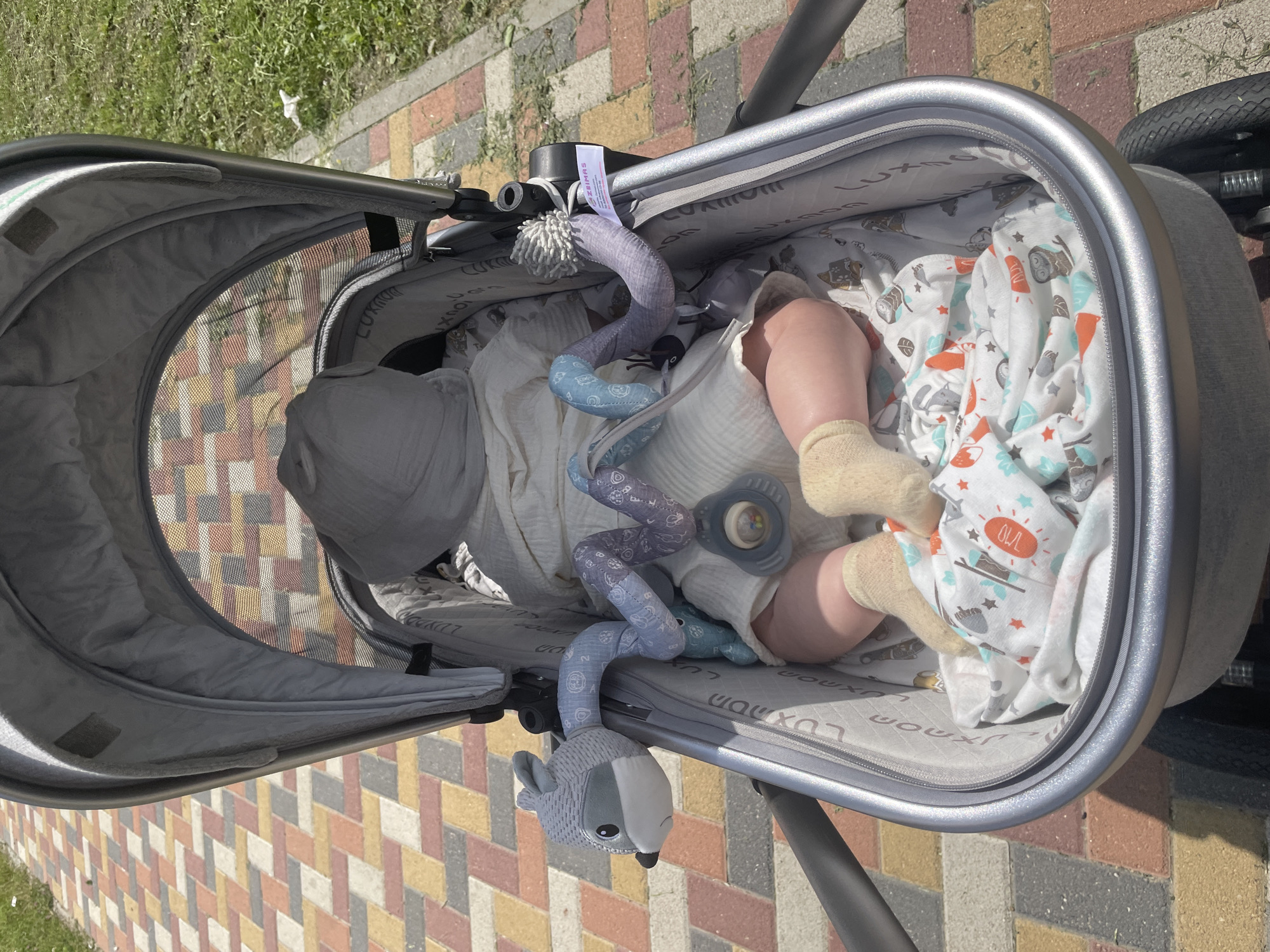 A baby is lying in a stroller on a paved path in Stavropol, Russia. The baby is covered with a light-colored blanket and wears socks. A toy is attached to the side of the stroller. The path is made of colorful bricks. Grass is visible on the right side of the image. The sky is clear and the sun is shining.